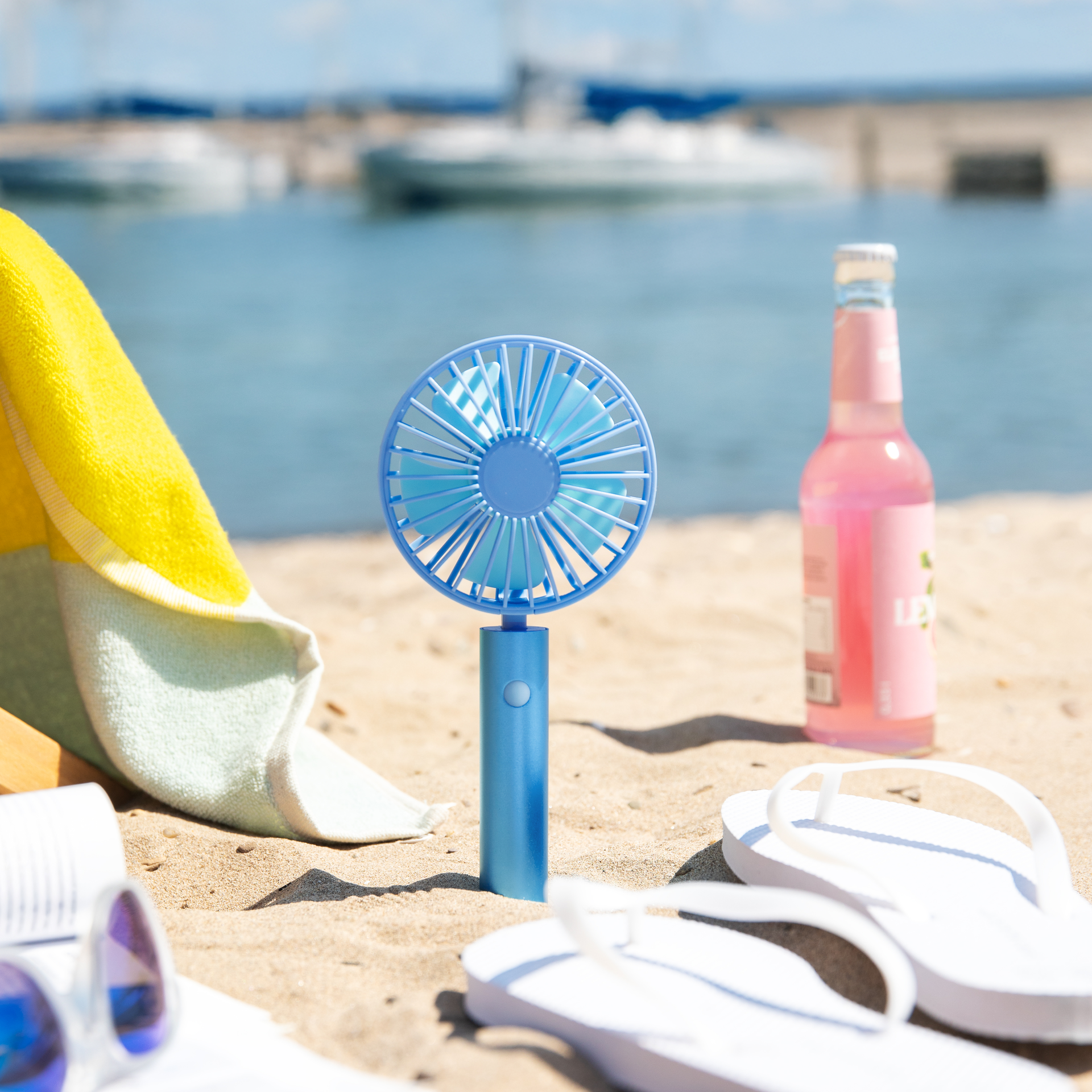 Ein blauer, aufladbarer Akku-Ventilator "Flow aqua" steht am Strand. Im Hintergrund sind Meer, Boote und ein Handtuch zu sehen. Flip-Flops und ein Getränk sind im Vordergrund.