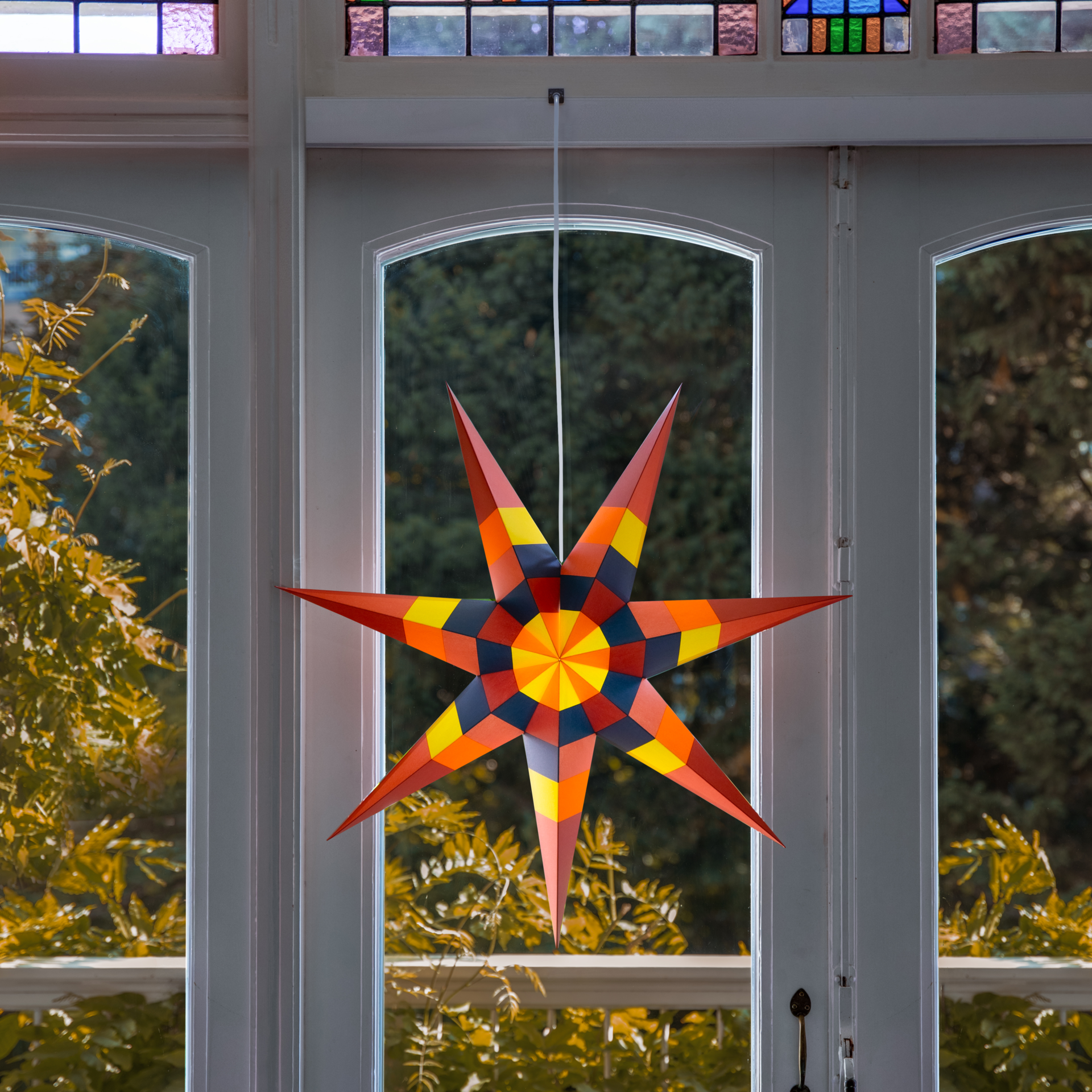 A colourful paper star in the window: a pattern of red, orange, yellow and dark blue rays, hanging from a white string. Blurred plants in the background.