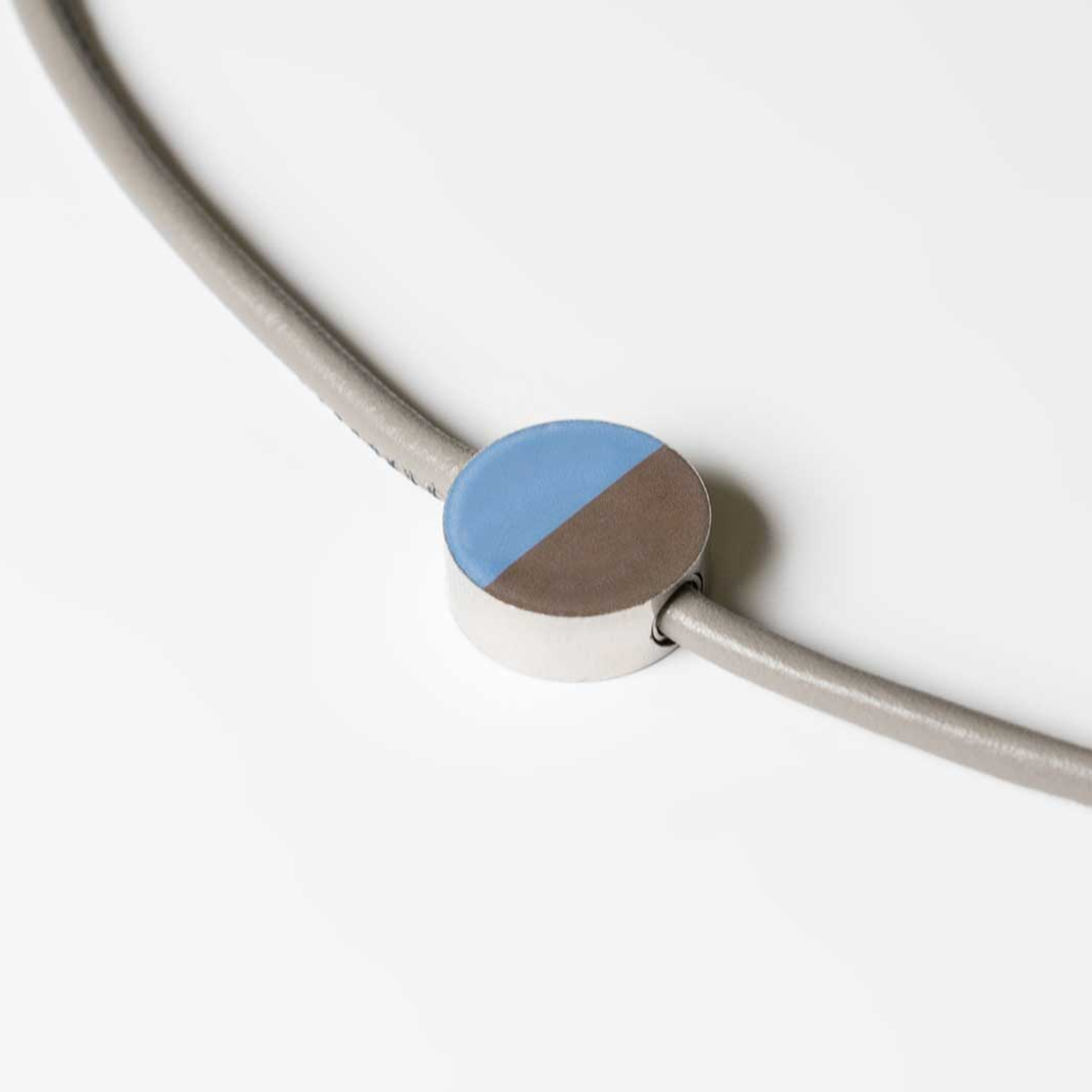 Primo piano del ciondolo gioiello "15" in acciaio, con una metà in blu e l'altra in marrone. Il ciondolo è fissato a un nastro color tortora.