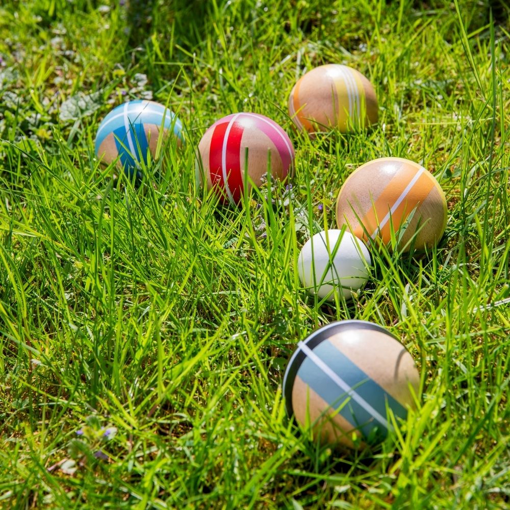 Mehrere bunte, gestreifte Holzbälle eines Rasenspiels liegen verstreut im frischen, grünen Gras unter hellem Sonnenlicht. Ein kleiner weißer Zielball befindet sich zentral zwischen den größeren Bällen, die in verschiedenen leuchtenden Farben bemalt sind.