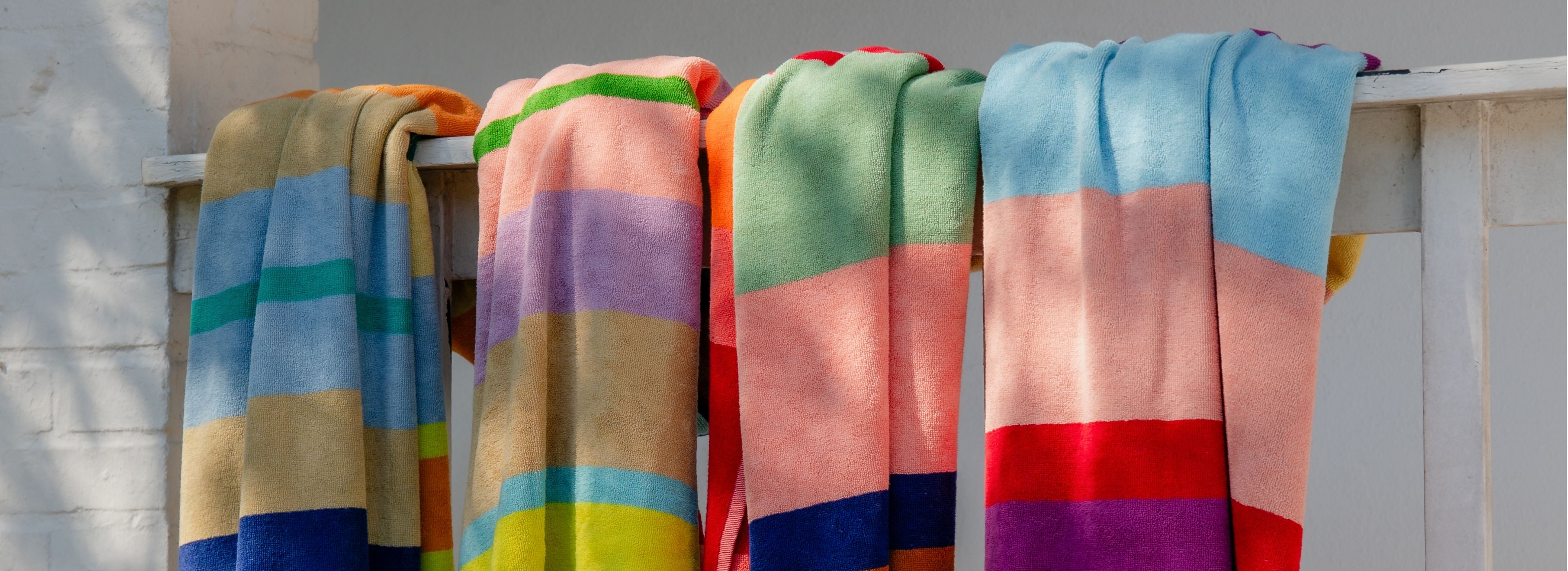 Vier bunte Handtücher mit breiten horizontalen Streifen hängen über einer weißen Holzstange. Sie zeigen Blau, Gelb, Türkis, Orange, Rosa, Lila, Grün und Rot und trocknen im Sonnenlicht, das Schatten wirft.