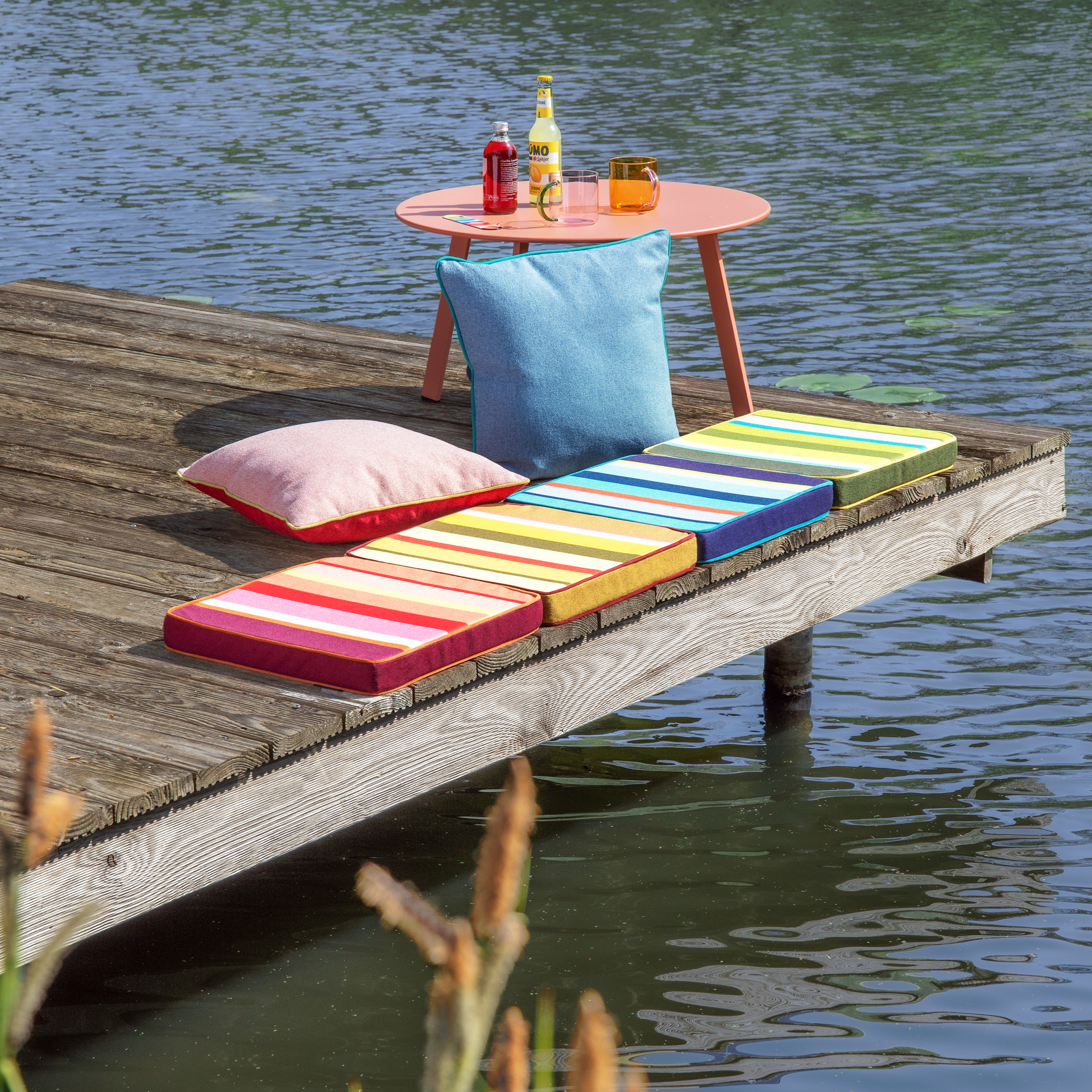 Passerelle en bois au bord de l'eau avec coussins colorés bleus et roses. Sur une petite table corail : boissons rafraîchissantes et verres. En arrière-plan, lac paisible avec nénuphars ; au premier plan, plantes aquatiques floues.