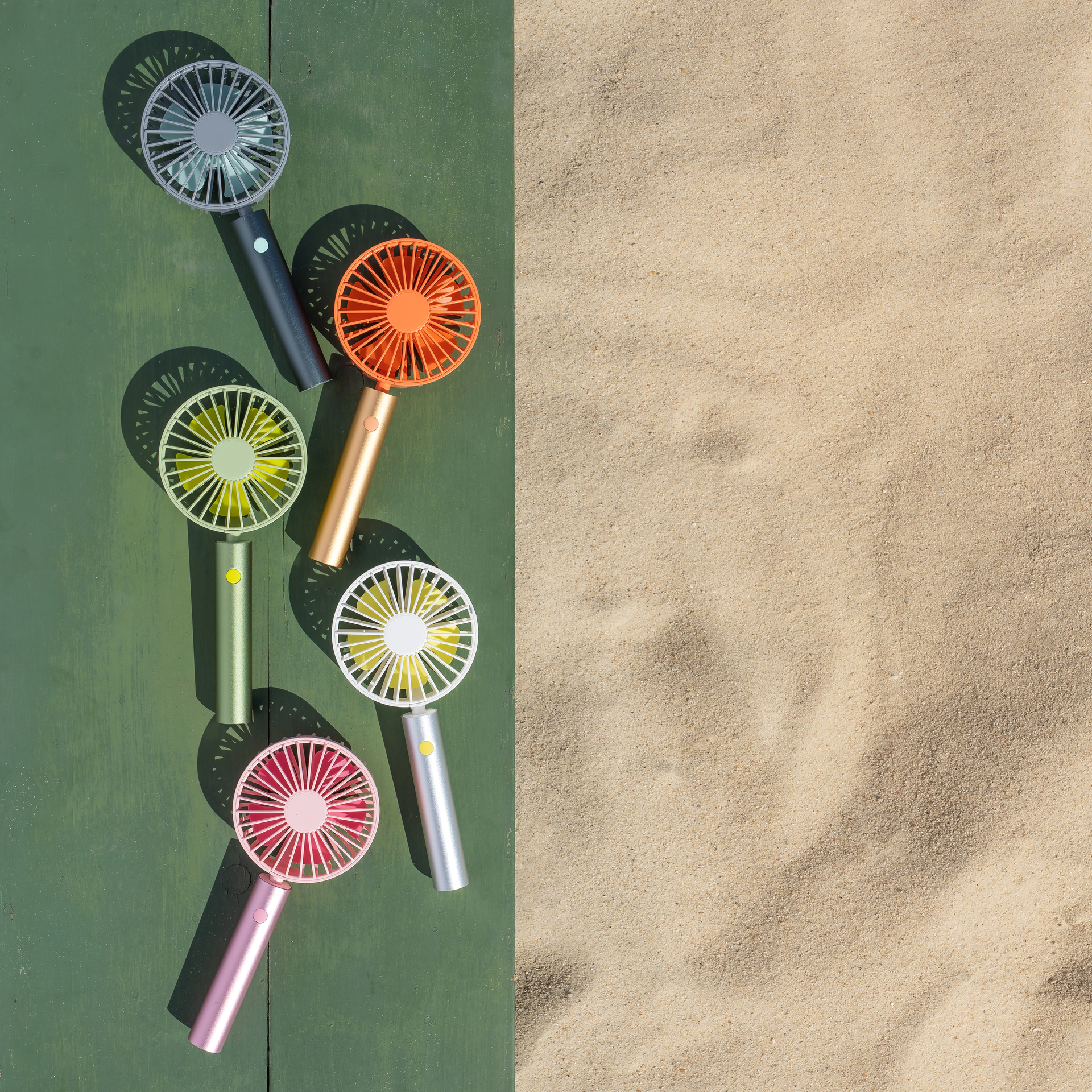 Ventilateurs Flow multicolores sur fond vert et sable, vue de dessus. Les ventilateurs sont disponibles en gris, orange, jaune, blanc et rose.