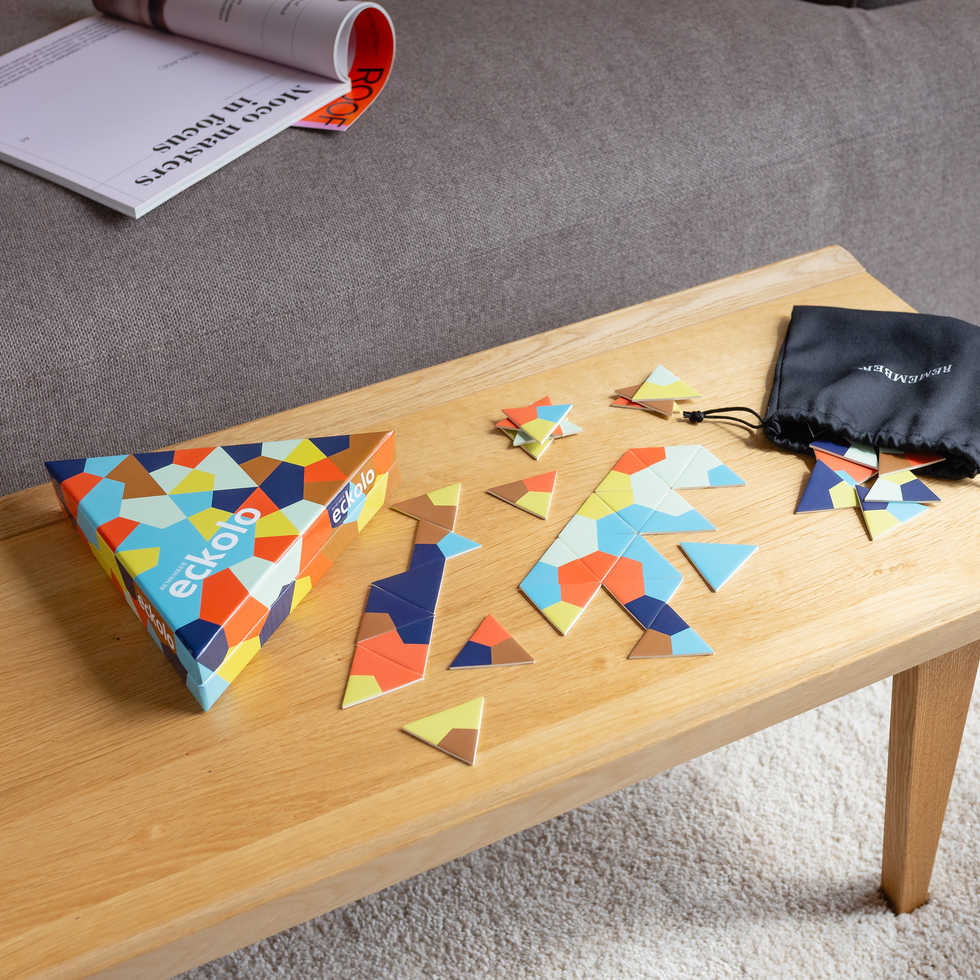 Eckolo liegt auf einem hellen Holztisch: dreieckige Box und viele dreieckige Karten mit farbenfrohen geometrischen Mustern. Daneben ein schwarzer Stoffbeutel mit weiteren Karten. Im Hintergrund ein graues Sofa.