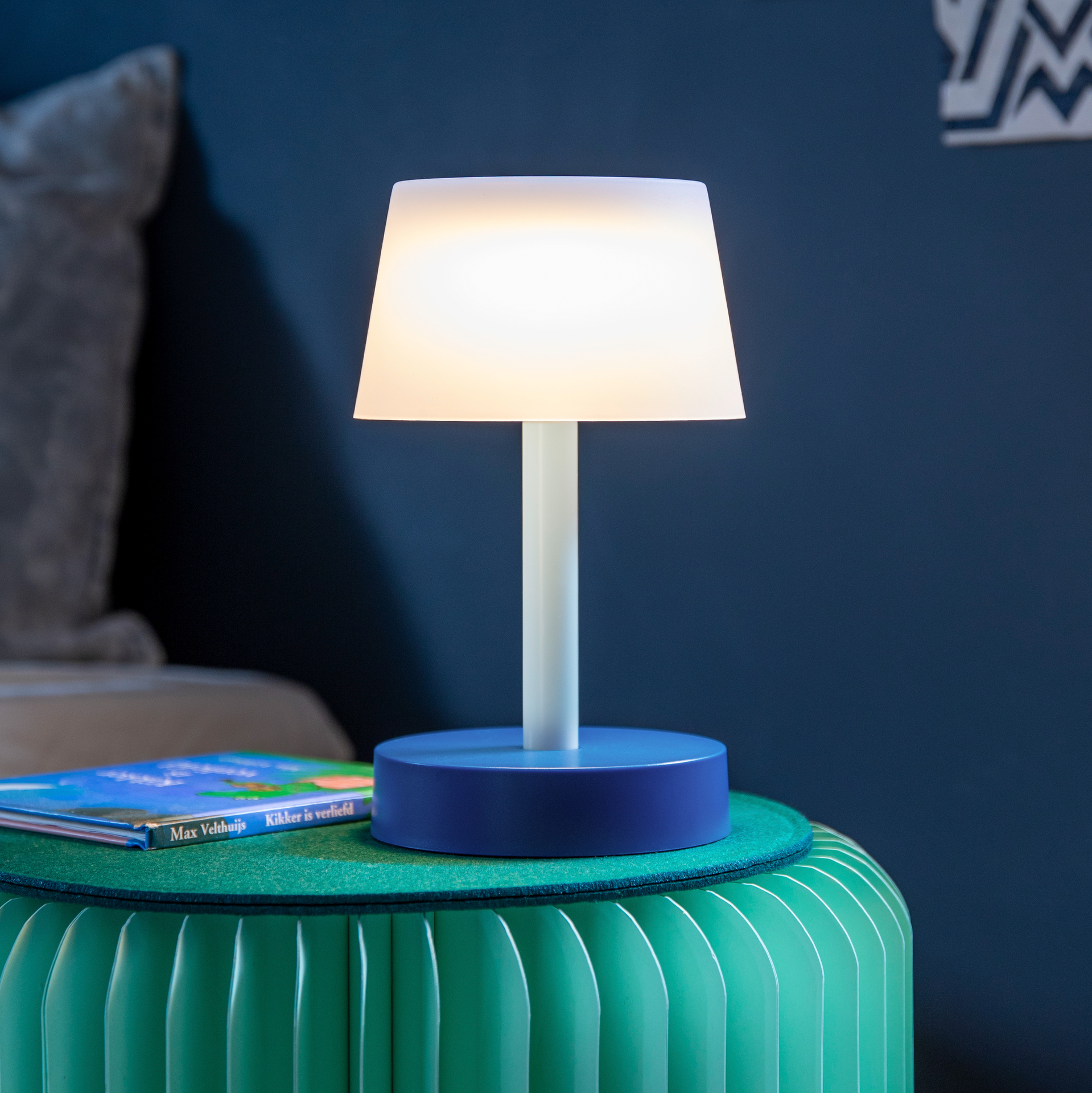 Een blauwe 'Fritz sky' tafellamp staat op een groen krukje. De lamp heeft een witte kap en straalt een warm licht uit. Er ligt een kinderboek naast.