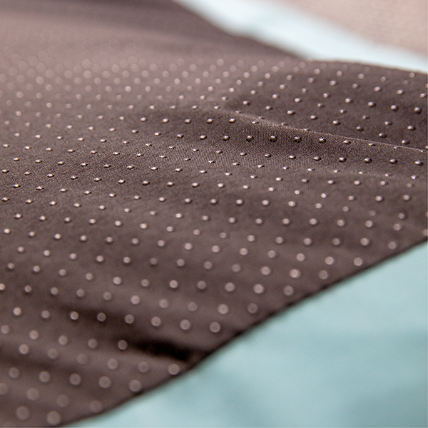 Dark brown fabric with numerous small, shiny, silver-coloured anti-slip dots, evenly distributed and visible as a textured pattern. Slightly pleated to create depth, with a blurred light blue area in the background.