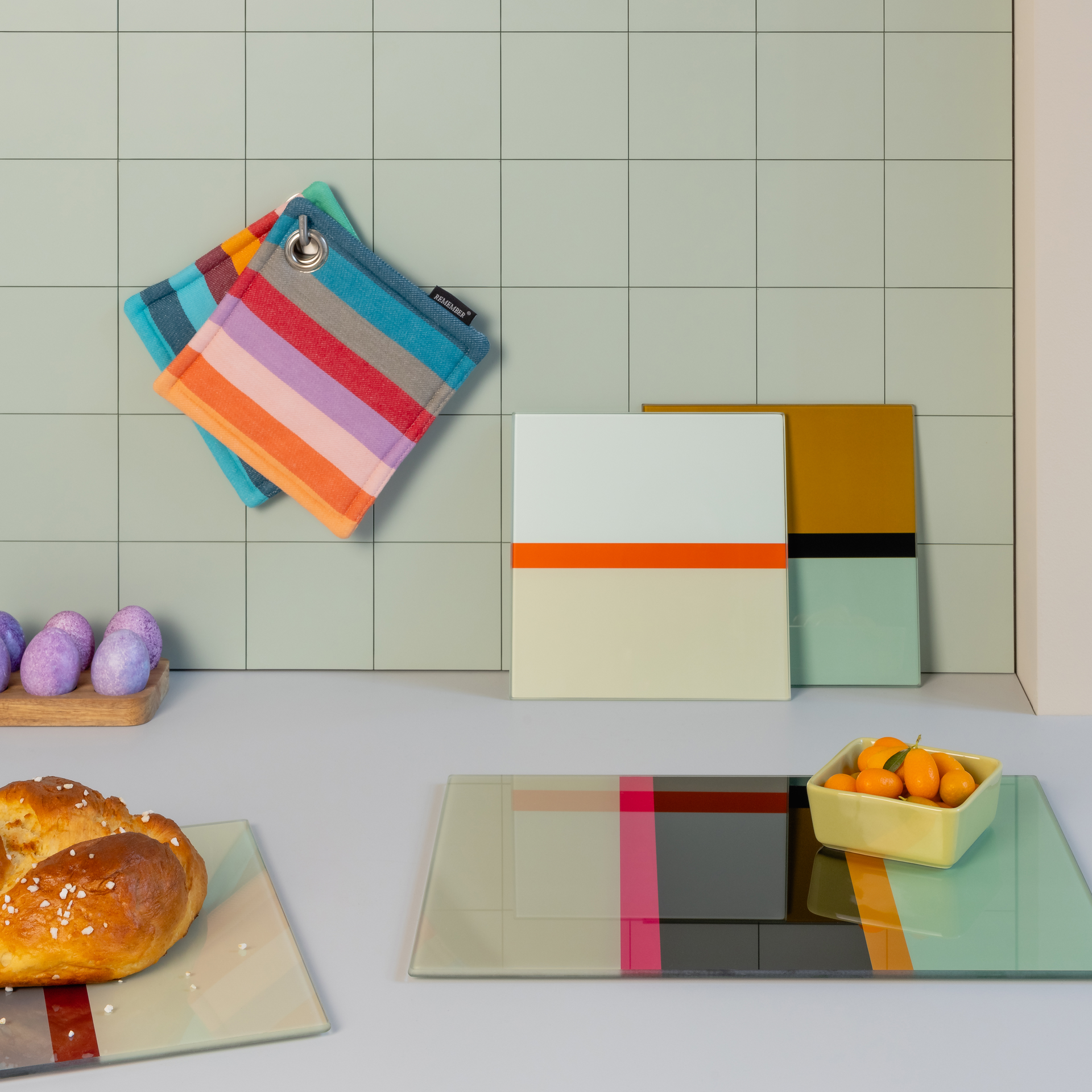Piani di vetro quadrati per la cucina con accenti colorati. Sullo sfondo, presine a righe, pane di Pasqua e kumquat in una ciotola.