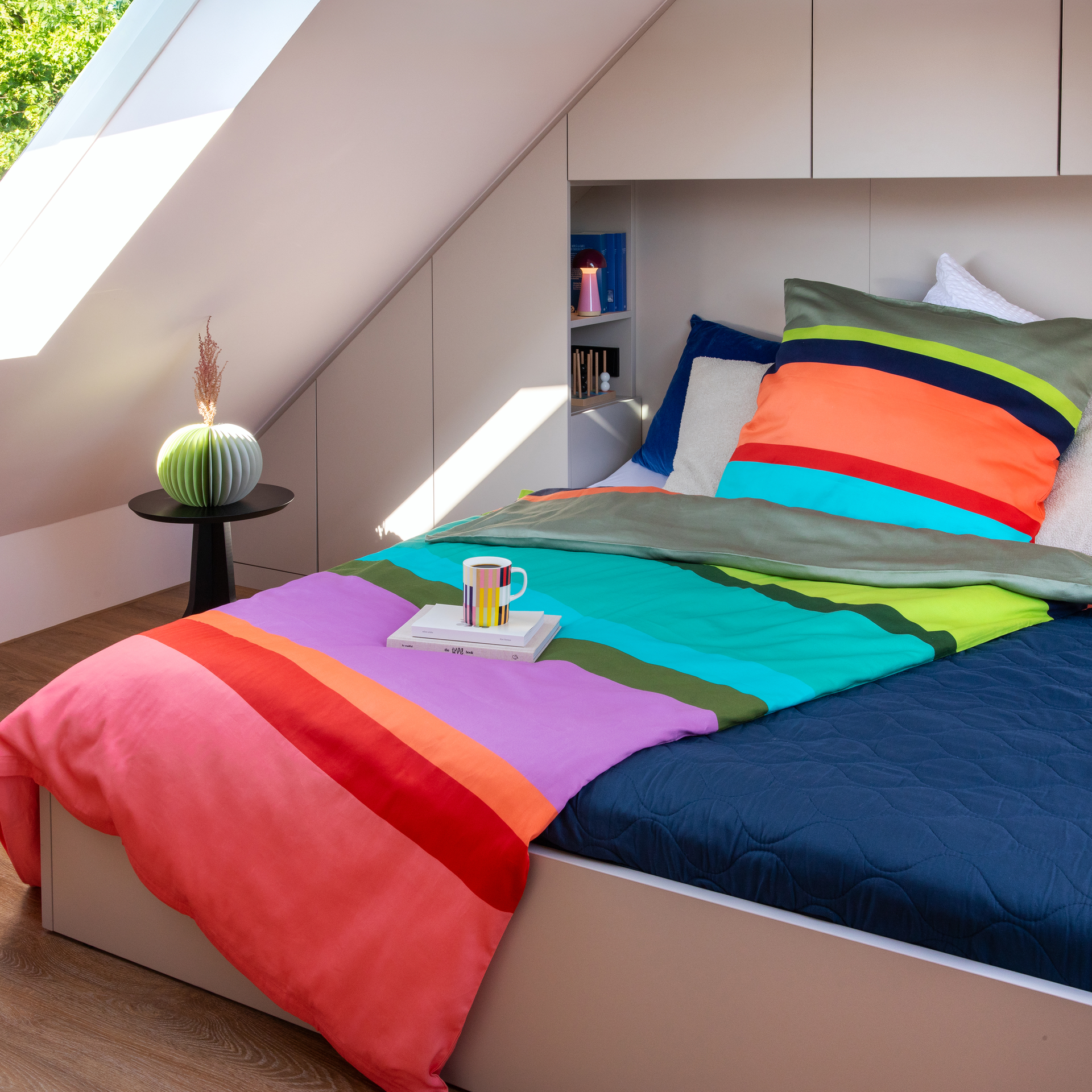 Gemachtes Bett mit Bettwäsche „Riva“ in leuchtenden Horizontalstreifen in Pink, Rot, Orange, Lila, Türkis und Grün. Auf der Decke liegen zwei Bücher und eine bunte Tasse. Im hellen Dachzimmer mit Seitenfenster und Sonne.