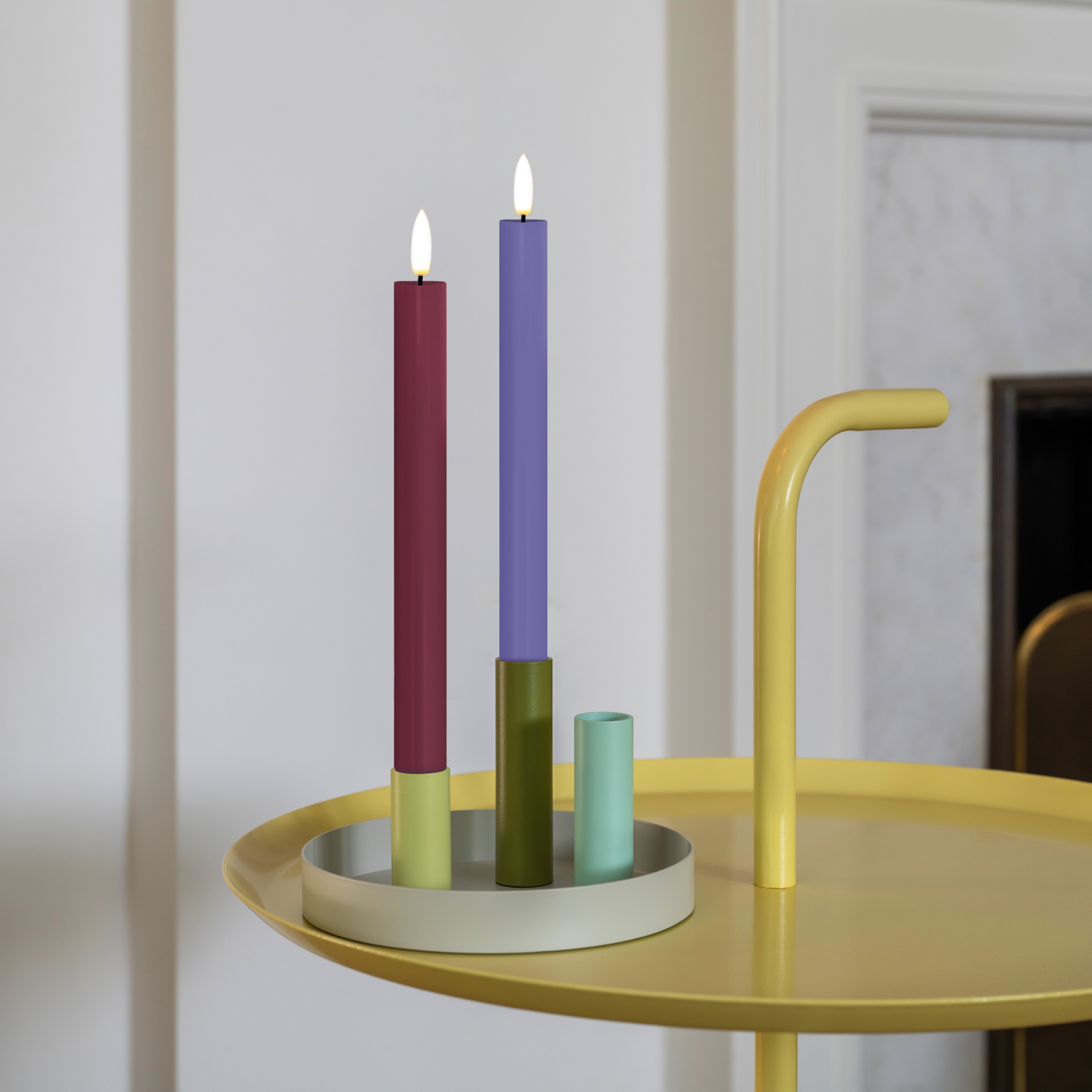Zwei LED Stabkerzen (bordeauxrot und lavendelfarben) auf weißem Hintergrund, mit weißem flammenförmigem Docht und geriffeltem weißem Sockel. Daneben eine weiße Fernbedienung mit Tasten. Höhe: 24,5 cm.