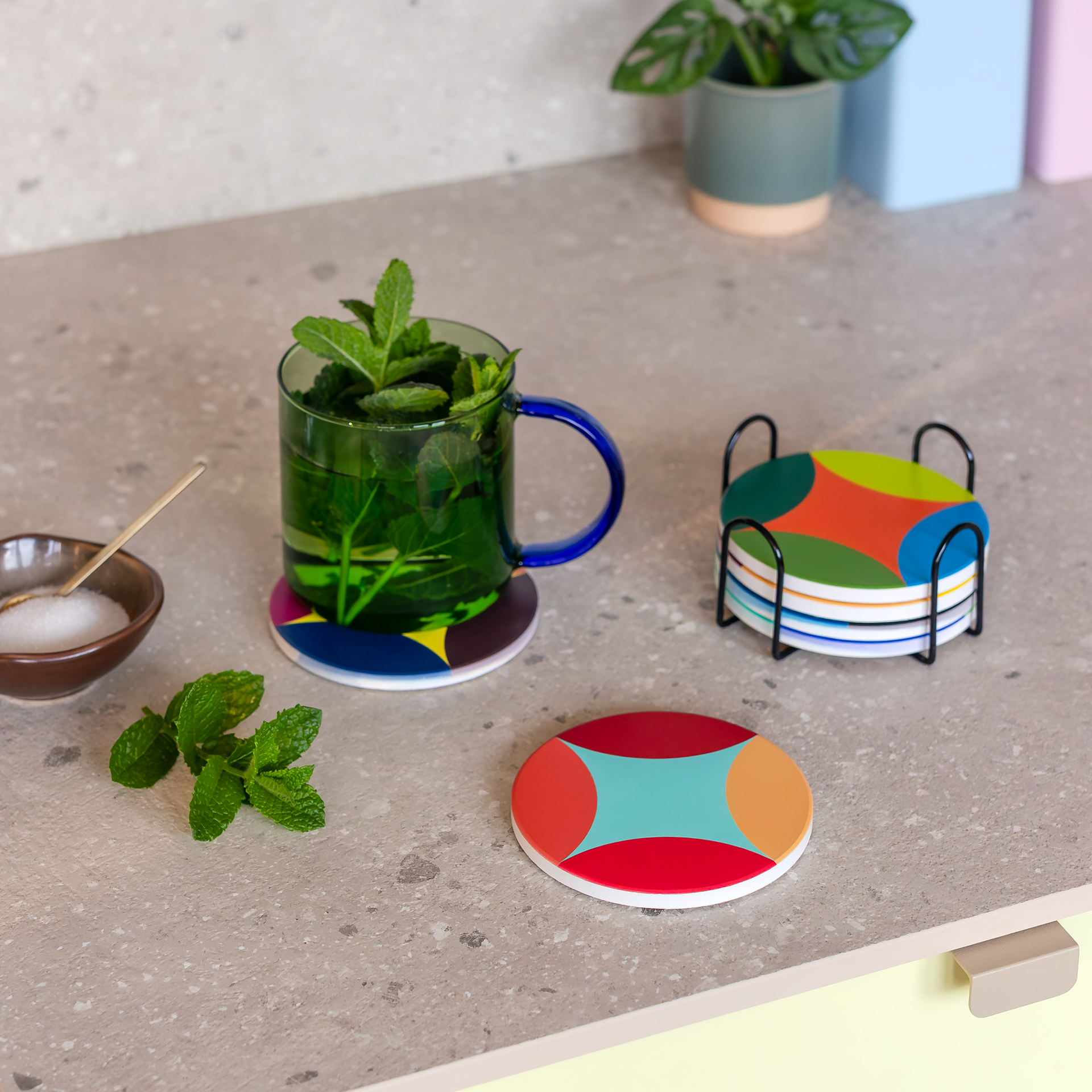 Six round, coloured ceramic coasters stacked in a black metal holder. Each has its own design, visible on the side and top. Top: a pattern of blue, orange, light and dark green areas. Cork underside, white rims.