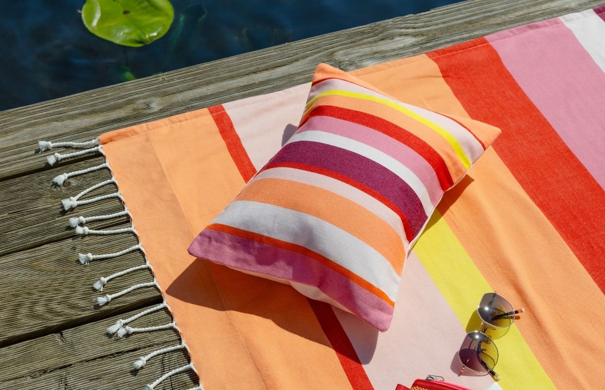 Ein gestreiftes Strandtuch in leuchtenden Orange-, Rot-, Pink- und Gelbtönen liegt auf einem Holzsteg am Wasser, daneben schwimmt ein grünes Seerosenblatt. Darauf: Kissen, Sonnenbrille und kleine rote Tasche. Entspannung am See.