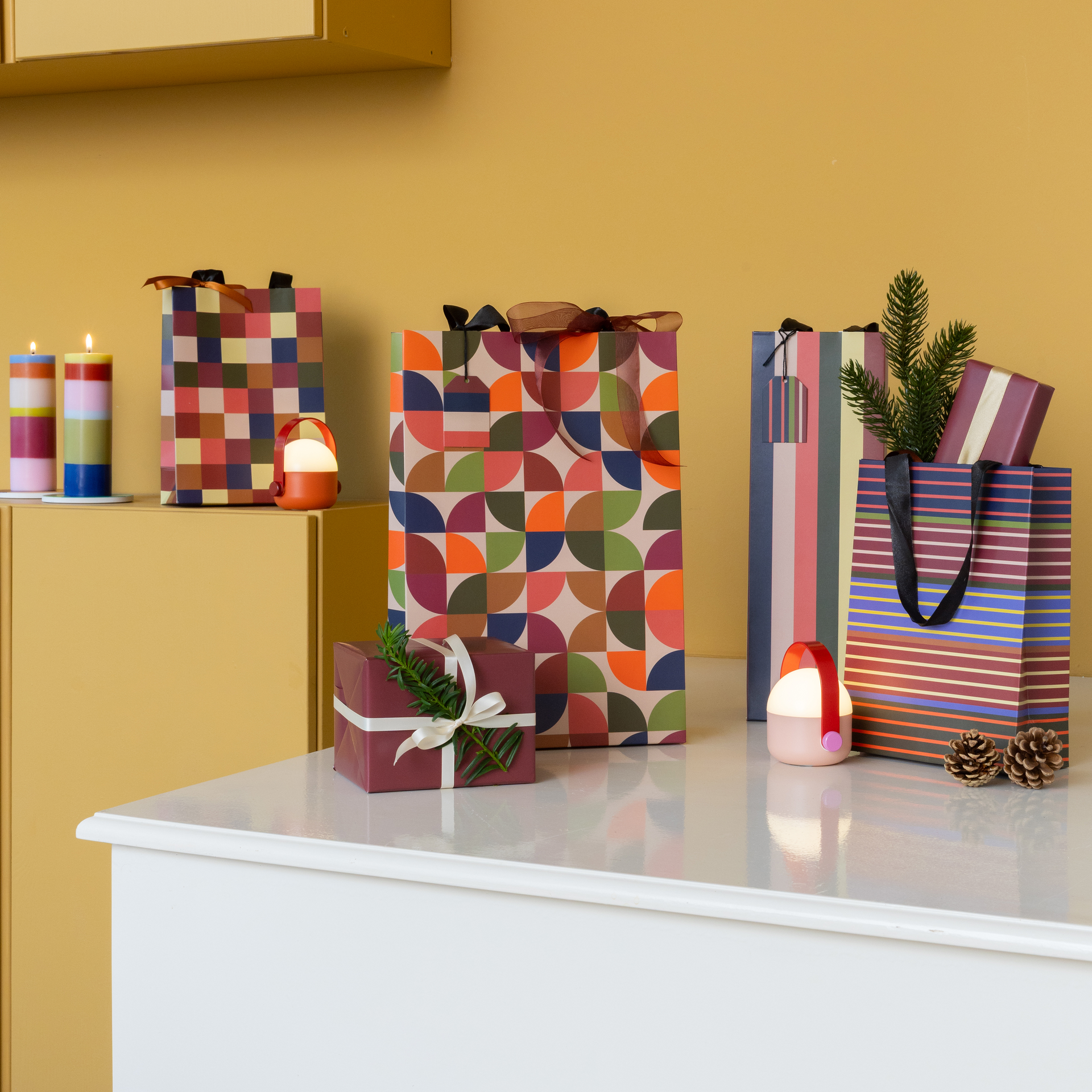 Verschillende kleurrijke cadeauzakjes met diverse patronen staan op een witte ondergrond. In één zakje zit een dennen takje. De feestelijke decoratie omvat ook ingepakte cadeautjes, brandende kaarsen en kleine lampjes tegen een gele achtergrond.