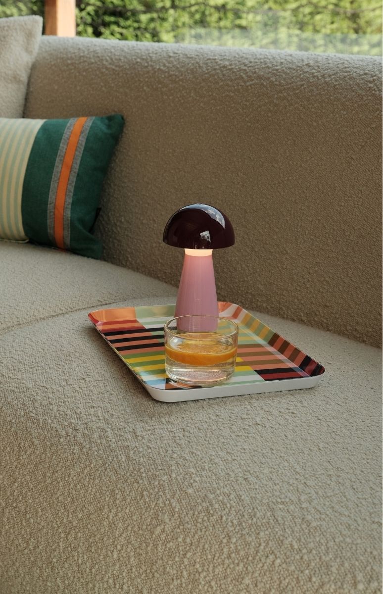 Gemütliche Szene auf beigem Sofa: pilzförmige Lampe mit dunklem Kopf und pinkem Fuß spendet mildes Licht. Daneben ein Glas Wasser mit Orangenscheibe auf bunt gestreiftem Tablett. Gestreiftes Kissen am Sofa.