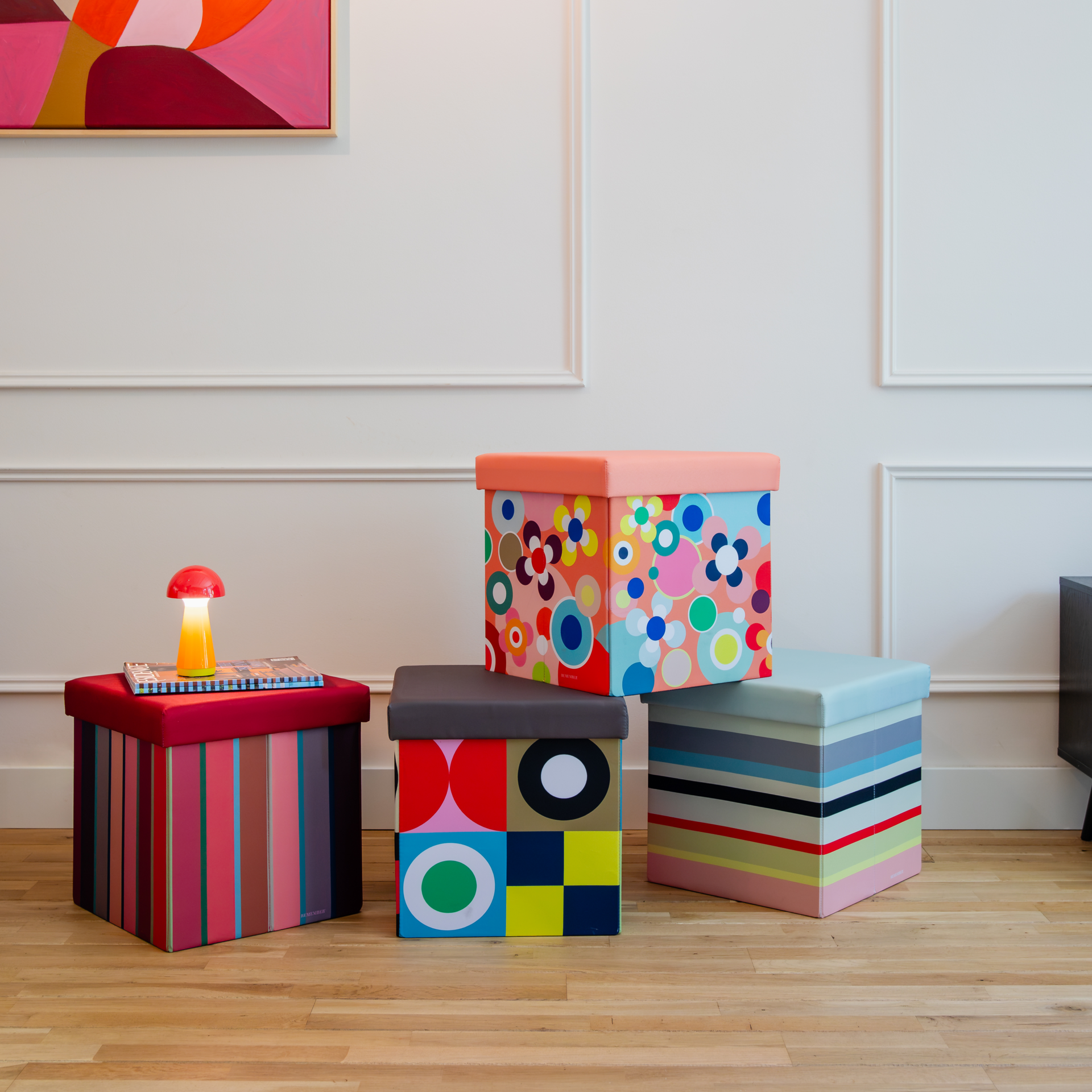 Vier bunte Sitzboxen mit Mustern auf hellem Holzboden vor weißer Wand. Links rot gestreift mit Pilzlampe und Magazinen. Mittig geometrisch, darüber floral. Rechts horizontal gestreift. Alle von Remember, als Stauraummöbel.