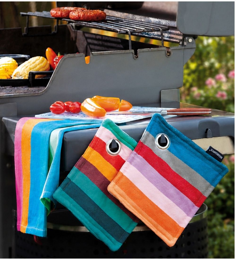 Ein Gasgrill steht im Freien, auf dessen Rost zwei Würstchen und Maiskolben garen. Auf der Ablage liegen frische Kirschtomaten und geschnittene orange Paprika. Ein blau-pink-oranges Tuch und zwei Topflappen mit Regenbogenstreifen hängen am Grill.