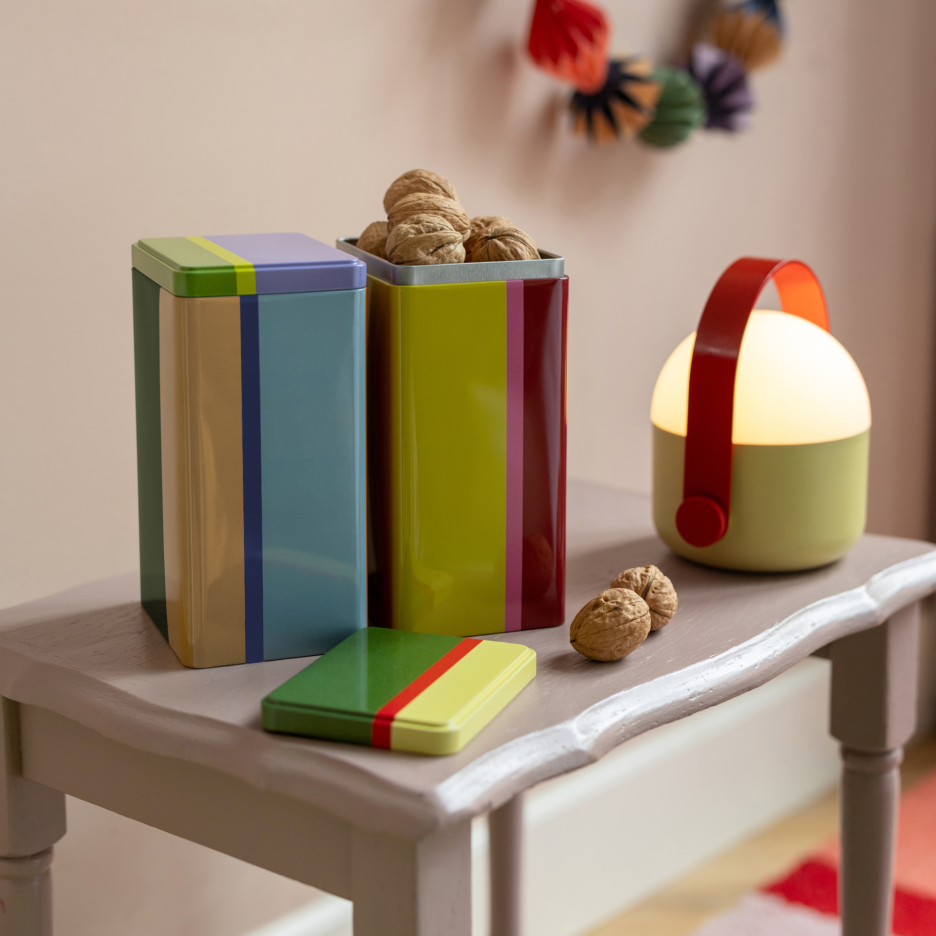 Farbiges Dosen-Set 'Yara' auf Tisch mit Nüssen und Lampe. Die Dosen sind gestreift in Blau, Gelb, Grün und Rot.