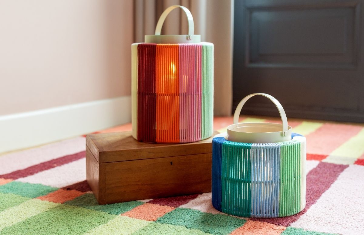 Zwei bunte, zylindrische Laternen stehen auf einem gemusterten Teppich. Die größere links steht auf einer Holzkiste, ist mit roten, orangenen, pinken und hellgrünen Fäden umwickelt, ihr Lichtschein ist sichtbar.