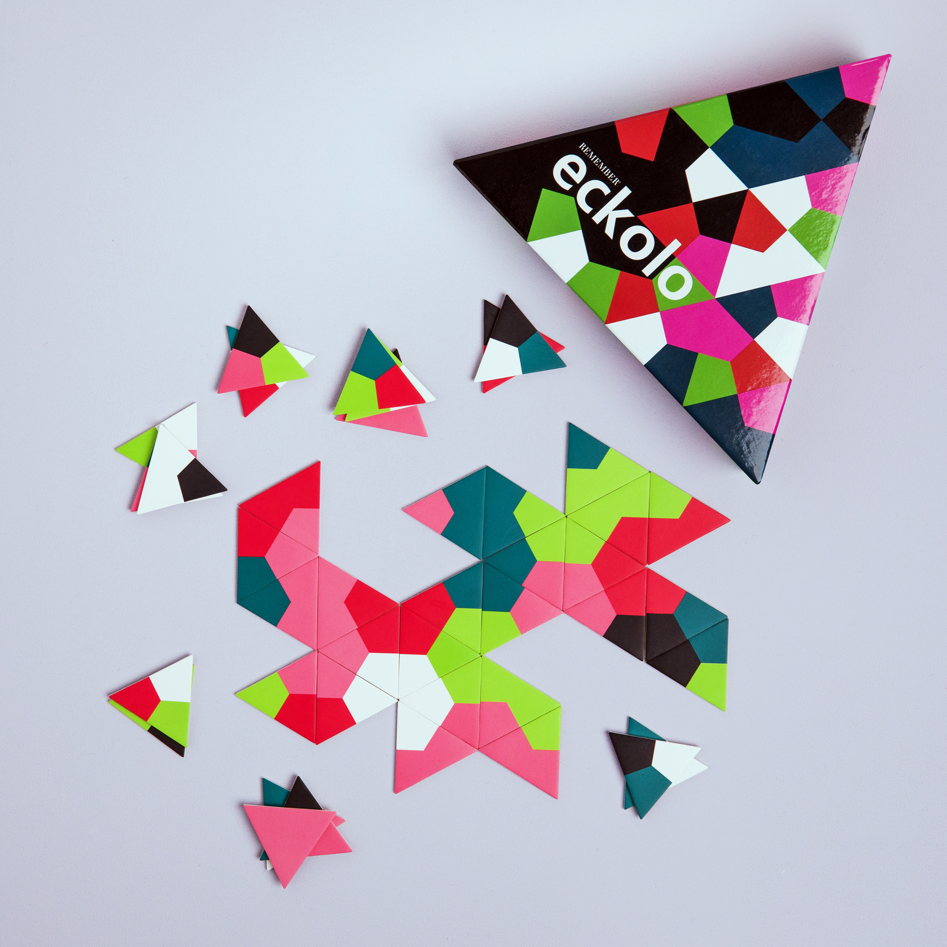 Il gioco di aggancio Eckolo: le carte da gioco triangolari di colore verde, rosa, rosso, nero e bianco si trovano in parte come una formazione a stella e accanto alla scatola di gioco triangolare su uno sfondo grigio.