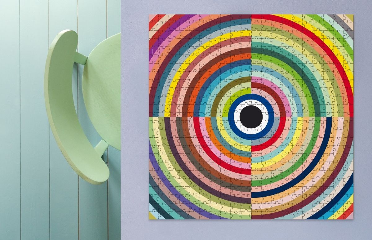 Quadratisches Puzzle mit farbigen konzentrischen Kreisen, ausgehend von einem schwarzen Punkt mit weiß-blauem Rand. Regenbogenringe in vier Quadranten. An grauer Wand; links Teil einer hellgrünen Stuhllehne vor blauen Holzpaneelen.
