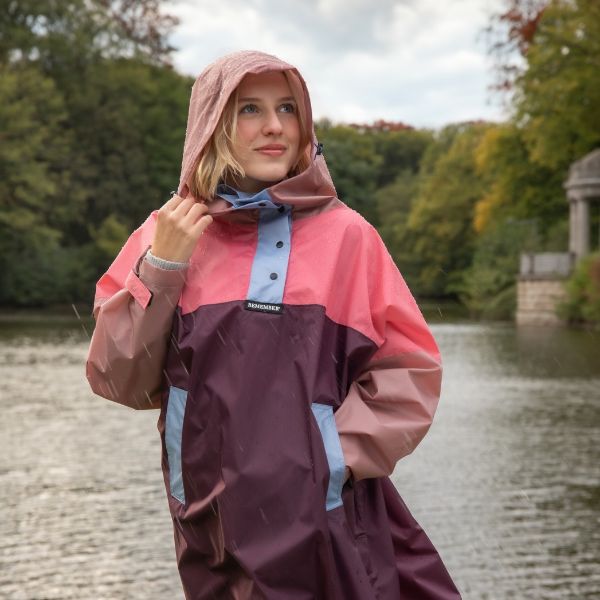 Eine junge blonde Frau trägt einen bunten Regenponcho in Rosa, Hellblau und Dunkelrot und zieht die Kapuze hoch. Regentropfen sind sichtbar. Sie lächelt in die Ferne über ein Gewässer, mit Bäumen im Hintergrund.