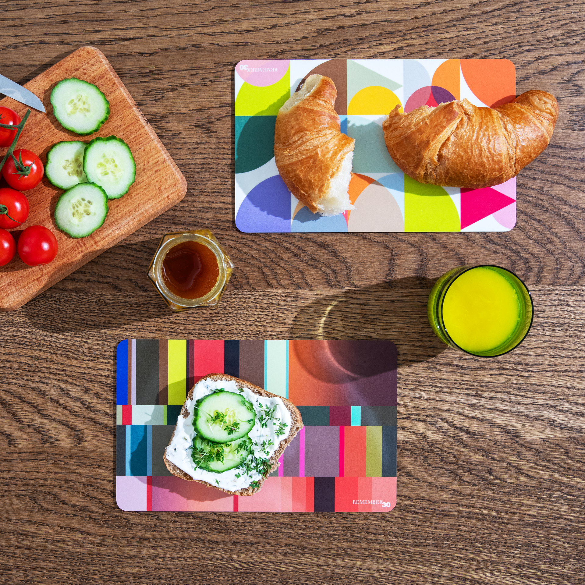 Draufsicht: Frühstück mit Croissants, Brot mit Gurke, Honig, Tomaten, Gurken auf Brett und Orangensaft auf buntem Untergrund.
