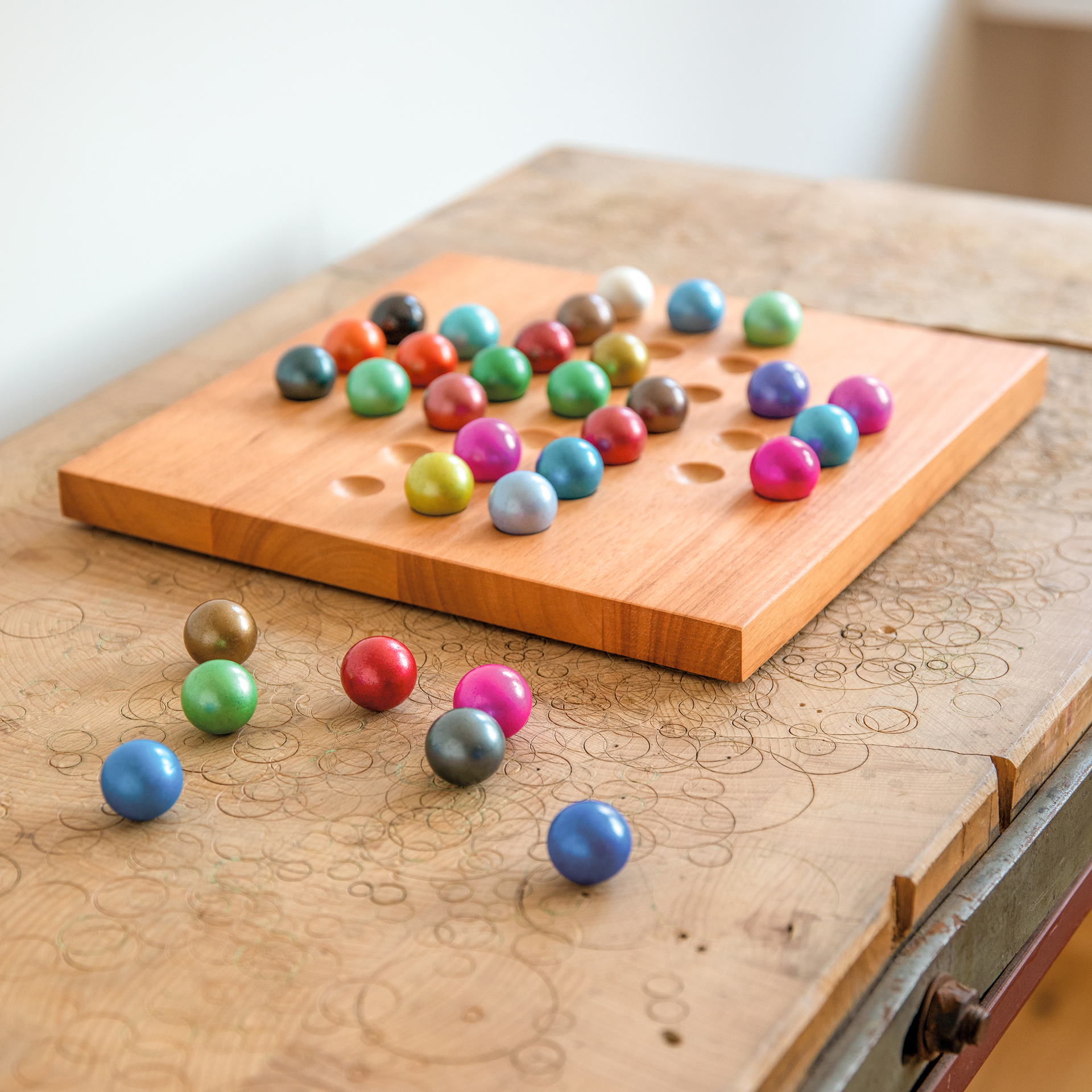 Wooden board with holes, filled with coloured glass balls. Some balls lie next to it. The solitaire game stands on a table with engravings.