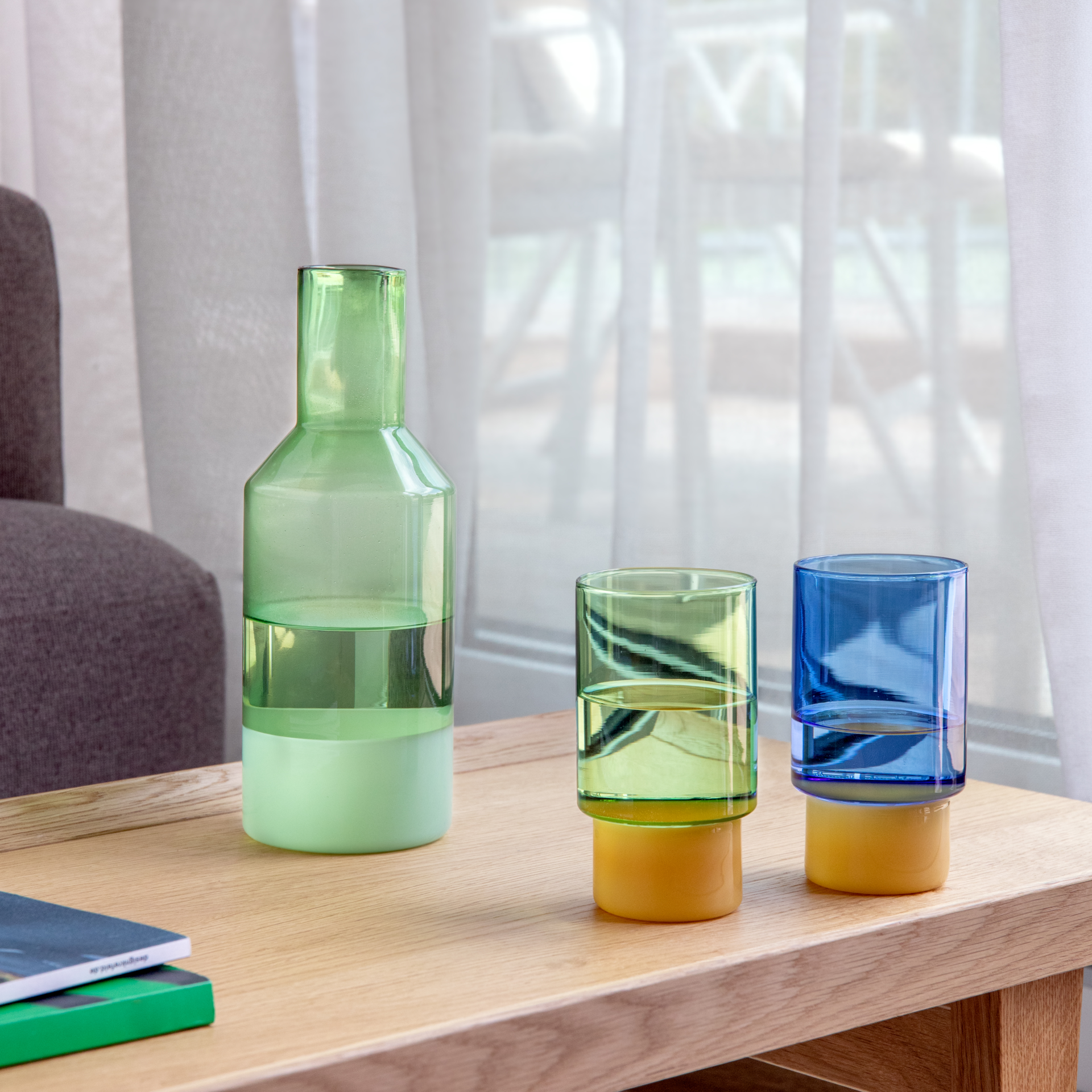 Drei stilvolle Gefäße aus Borosilikatglas auf hellem Holztisch: links grüne Karaffe mit Wasser, daneben grünes Jill-Trinkglas mit gelbem Sockel. Rechts blaues Jill-Trinkglas mit gelbem Sockel, ebenfalls mit Wasser. Helle Gardinen, unscharfes Fenster.