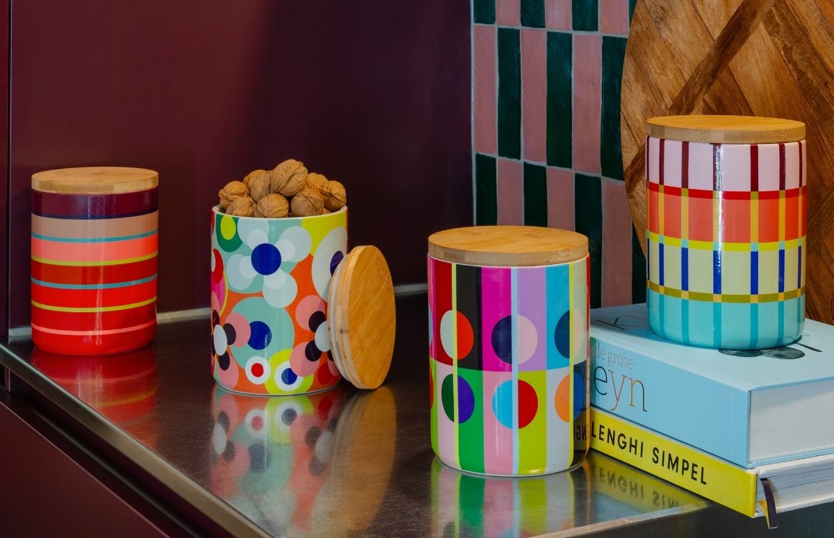 Vier bunte Keramikdosen mit Holzdeckeln auf glänzender Fläche; eine offen mit Walnüssen. Muster: Streifen, Blumen, Karos. Eine steht auf Büchern. Dunkelrote Wand im Hintergrund.
