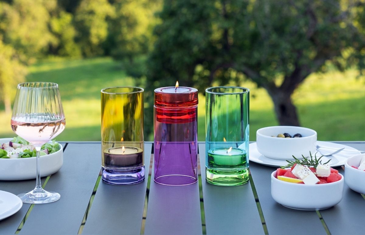 Gedeckter Tisch im Freien bei Tageslicht. Auf grauer Tischplatte: Glas Roséwein, Salatschüssel, Schale mit Oliven sowie Wassermelone und Feta. Gelbe, rote und grüne Glaszylinder mit Teelichtern.