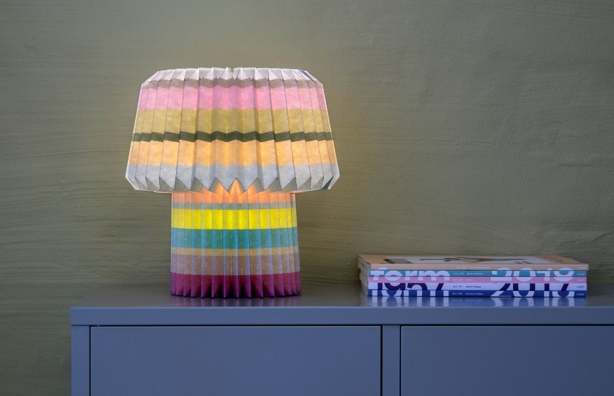 Leuchtende Tischlampe mit plissiertem Schirm und Fuß, horizontal gestreift in Rosa, Gelb, Grün, Blau und Grau. Sie steht auf einem schiefergrauen Schrank neben Magazinen vor olivgrüner, strukturierter Wand. Warmes Licht.