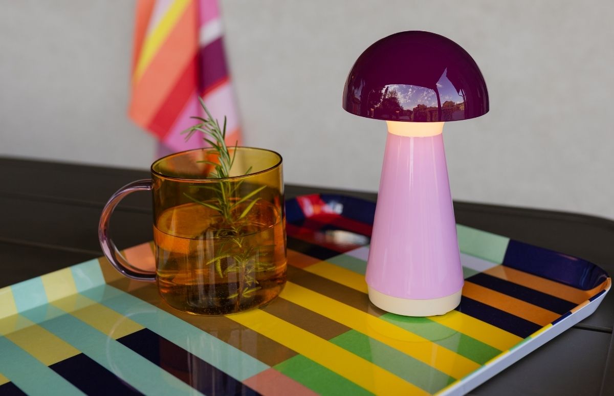 Violette Pilzlampe mit hellrosa Ständer auf buntem gestreiften Tablett. Daneben bernsteinfarbene Glastasse mit lila Henkel, gefüllt mit Wasser und Rosmarinzweig. Gelb-Grün-Blau-Orange Streifen. Im Hintergrund gestreiftes Tuch an der Wand.