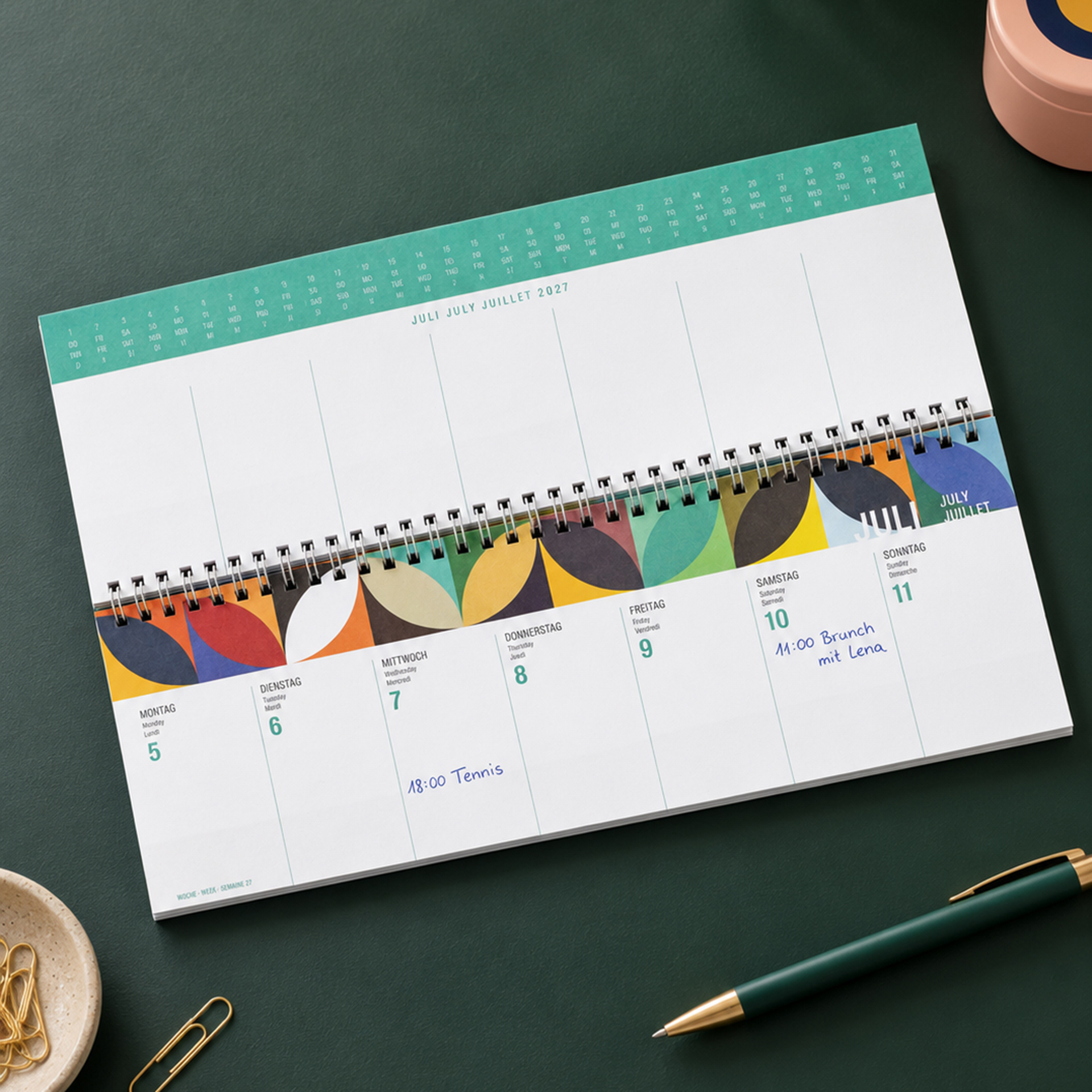 Agenda hebdomadaire 2027 ouvert en juillet sur bureau vert foncé ; semaine du 5 au 11 avec rendez-vous manuscrits « 18 h : tennis » et « 11 h : brunch ». Reliure spirale visible, graphisme en haut, stylo et trombones à côté.