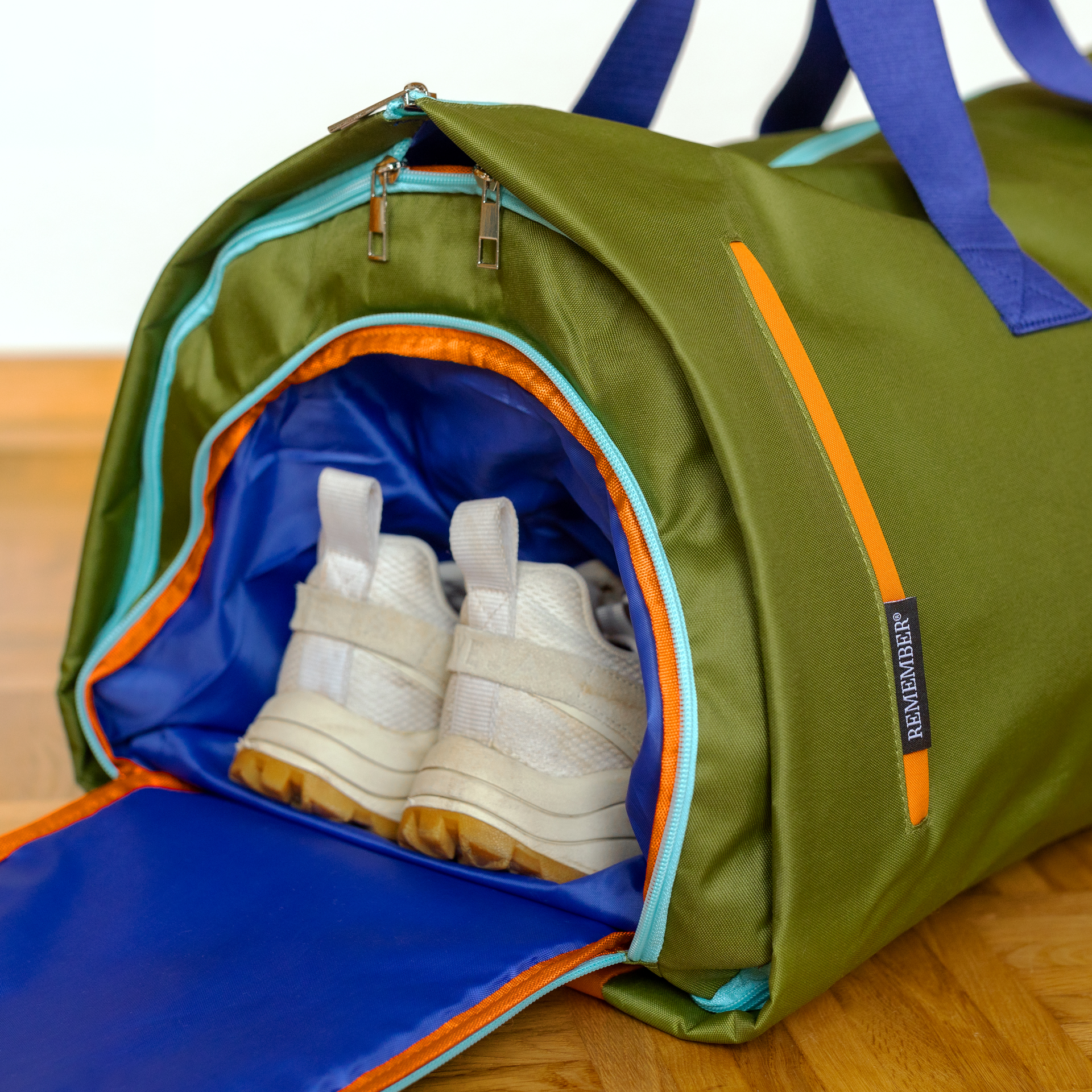 A partially open Lennard travel bag in dark forest green on a wooden floor. In the bright blue inner compartment of the side shoe compartment lie white trainers with light brown soles. Light blue zips, orange trim, ‘ REMEMBER’® label.