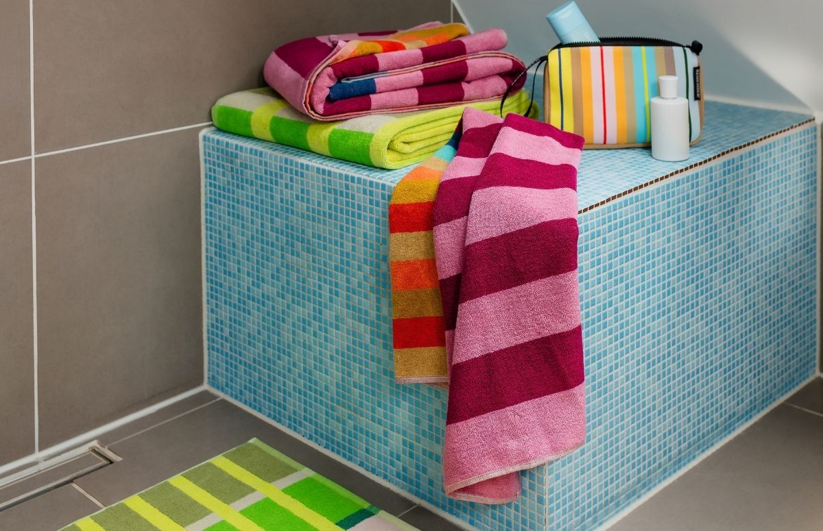 Bunte, gestreifte Handtücher liegen und hängen auf einem Badregal vor hellblauen Mosaikfliesen. Pink-lila und gelb-grüne Tücher sind gefaltet, orange-gelbe und pink-weinrote hängen herab. Daneben Kulturtasche und weiße Flasche. Grüne Badematte.