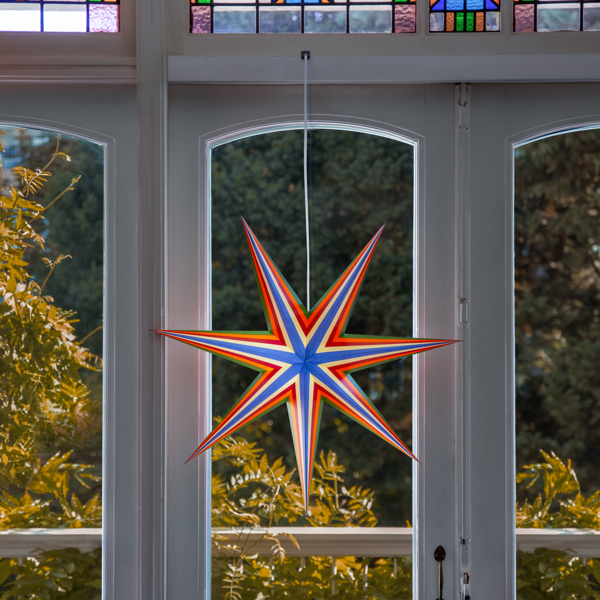 The colourful paper star ‘Mira’ hangs in front of a white window front. It has several points and stripes in red, orange, yellow, green and blue. Outside, you can see trees with yellow-green foliage. Above them are narrow stained-glass windows.