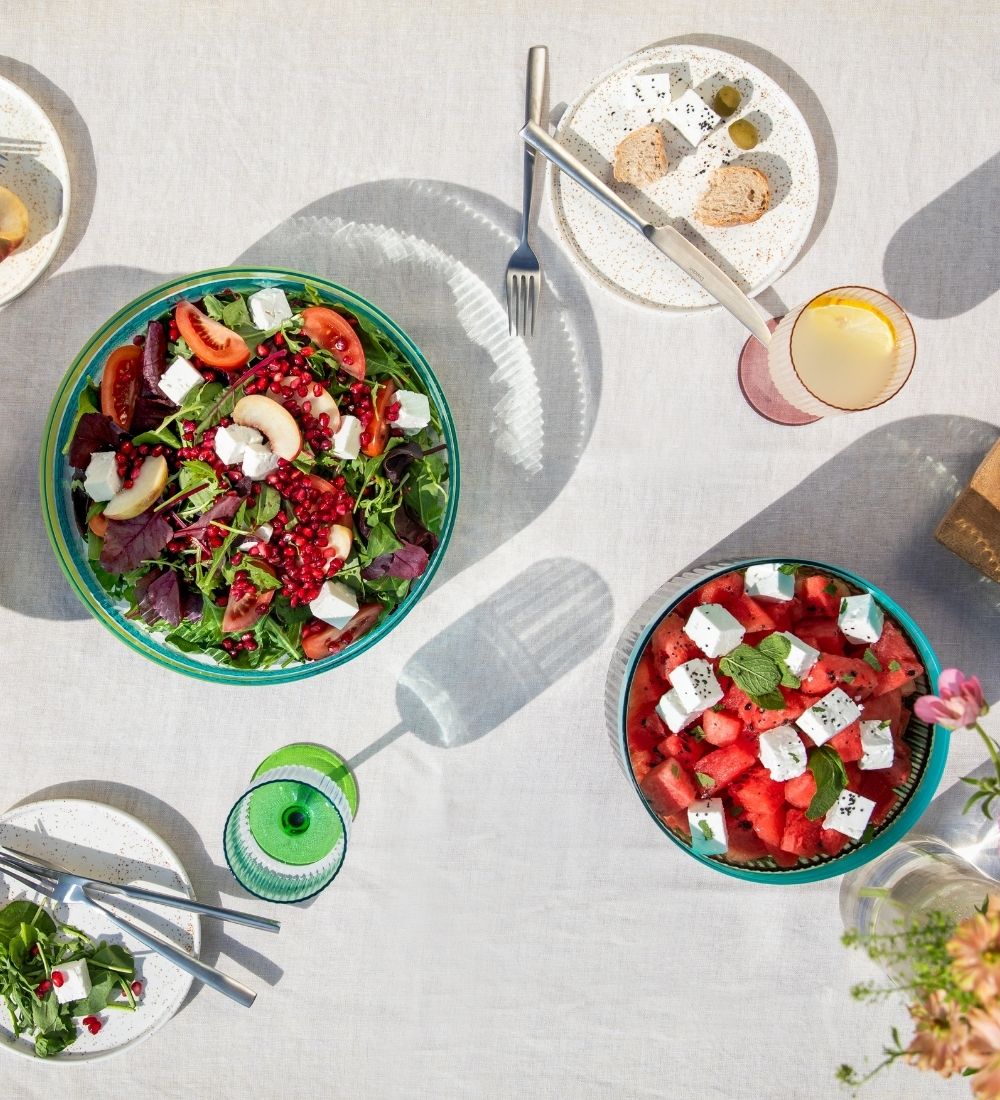 Sommerlich gedeckter Tisch im Freien, von oben. Zwei Salatschüsseln im Mittelpunkt: Blattsalat mit Tomaten, Feta, Birnen, Granatapfel; Wassermelone mit Feta und Minze. Teller, Besteck, Gläser, Brot auf weißer Decke, starke Sonne wirft lange Schatten.