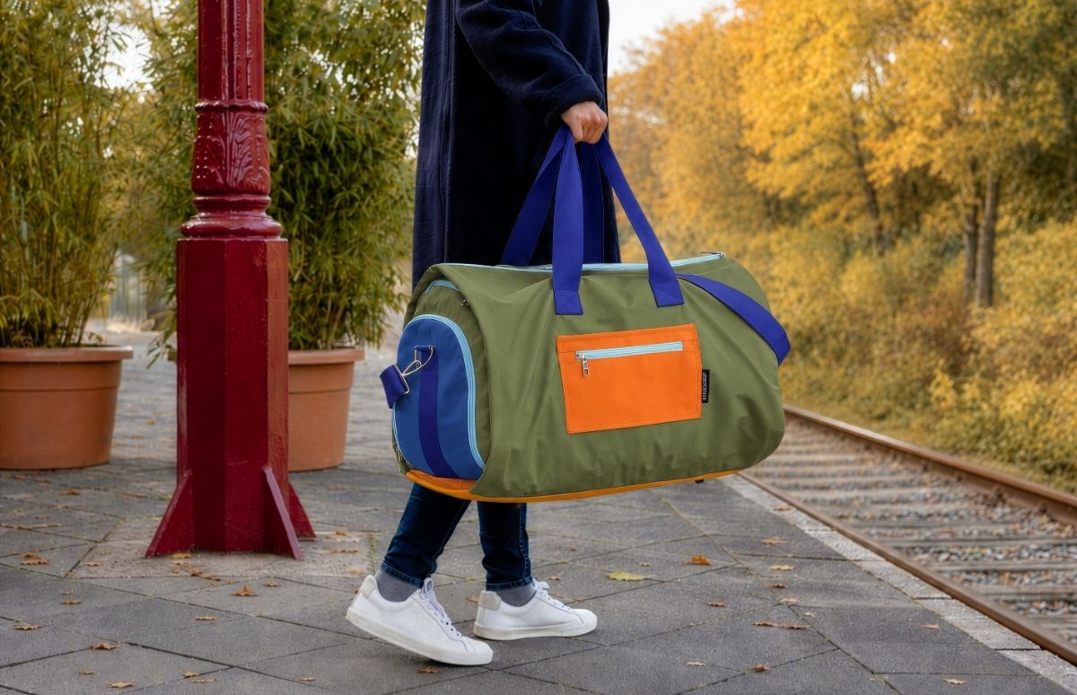 Person in dunklem Mantel, Jeans und weißen Sneakern trägt olivgrüne Reisetasche mit blauen Griffen, orangefarbener Fronttasche und blauer Seitentasche. Sie geht auf Bahnsteig mit Herbstlaub, neben rotem Pfosten.