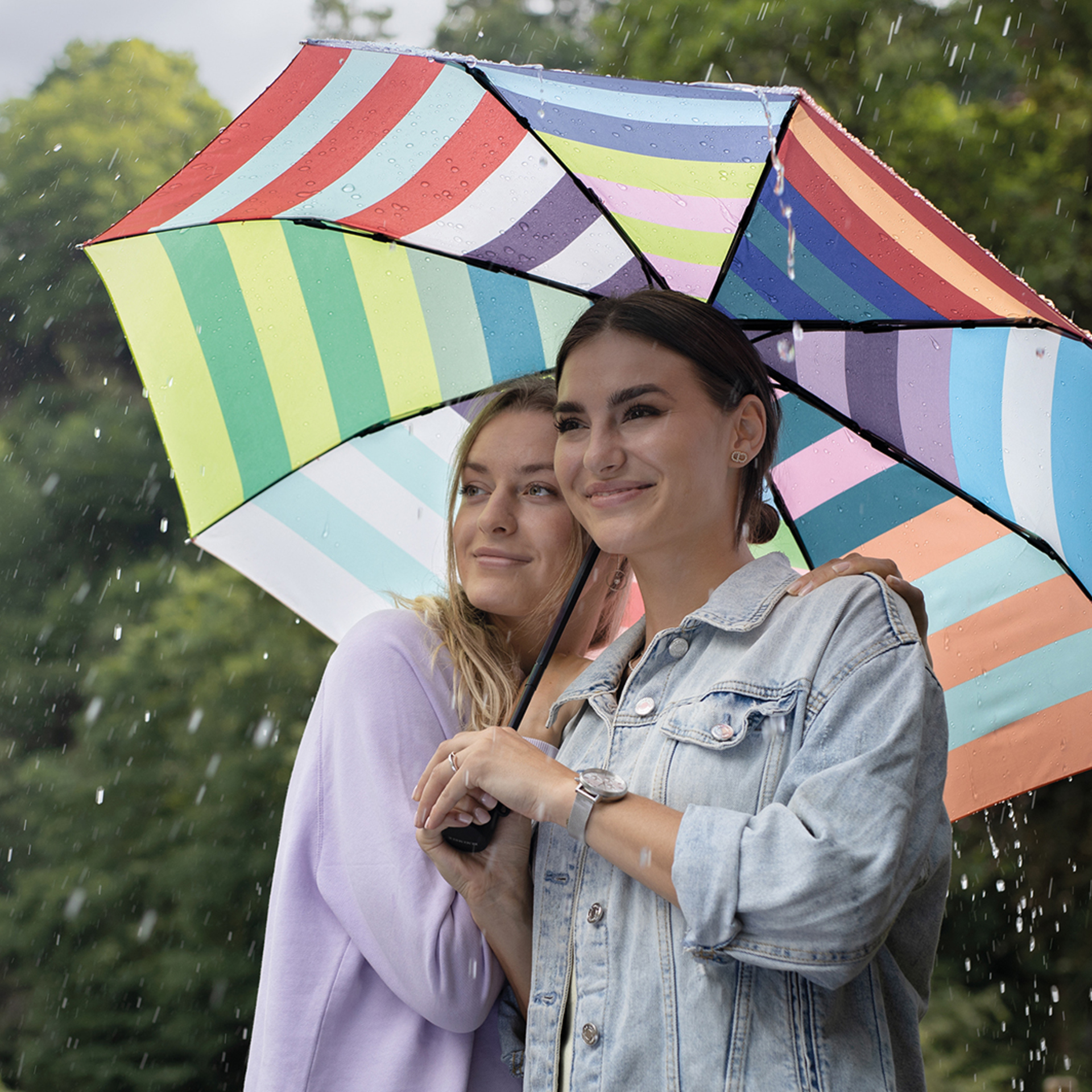 Two women under an open, colourful pocket umbrella in the rain. The umbrella has a striped pattern in different colours.