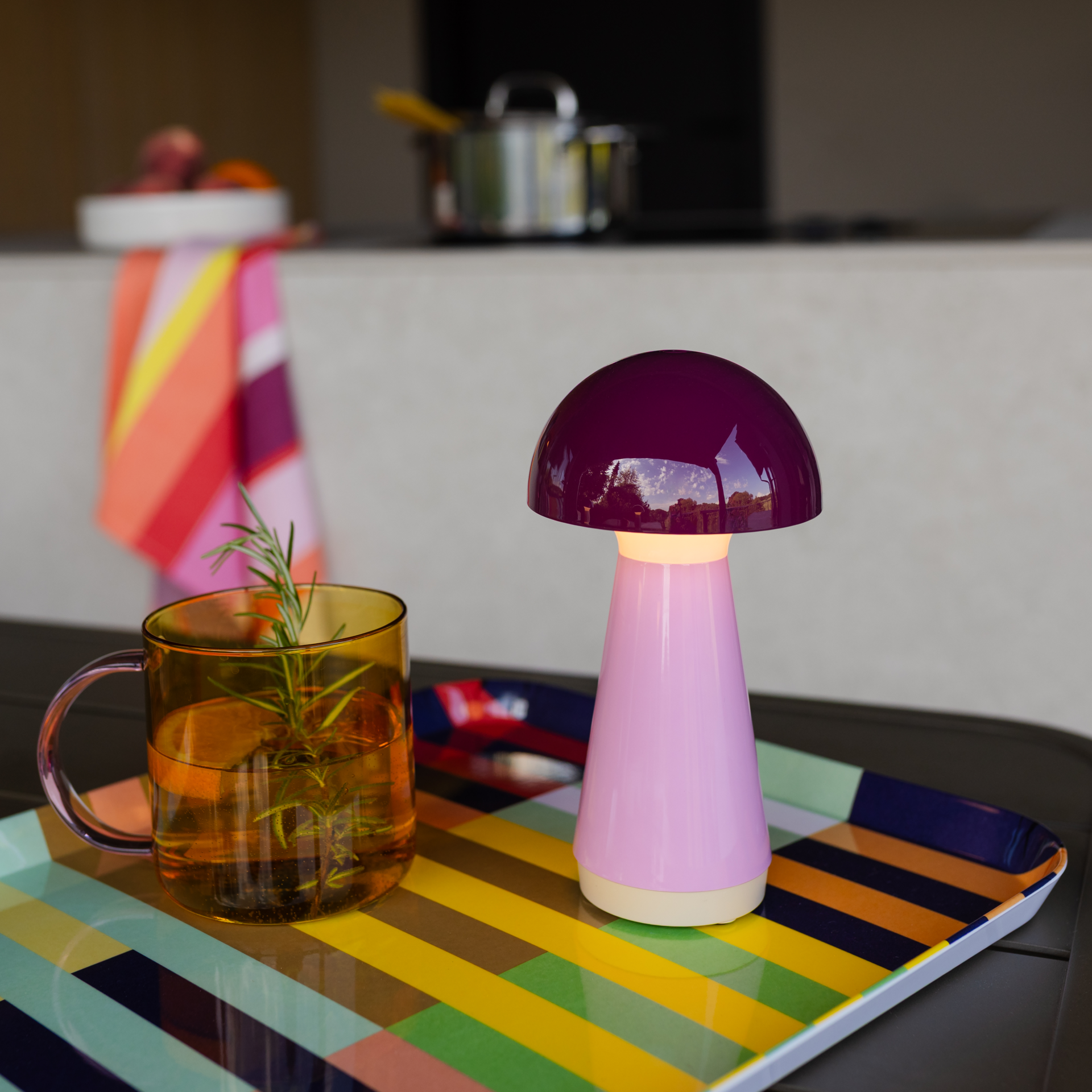La lampe de table Bobbi Berry allumée repose sur un plateau coloré à rayures. Abat-jour champignon violet foncé brillant, pied conique rose clair, lumière chaude. À côté, un verre ambré avec romarin. Extérieur en arrière-plan.