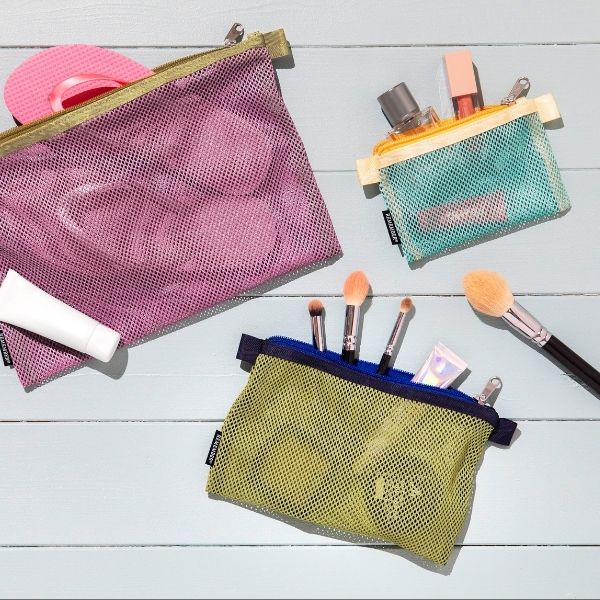 Drei farbige Mesh-Beutel mit Reiseartikeln und Kosmetika liegen auf hellblauem Holzhintergrund. Im violetten Beutel: pinke Flip-Flops und eine Tube Sonnencreme. Im türkisfarbenen: Parfüm und Lipgloss. Im limettengrünen: Make-up-Pinsel und Creme.