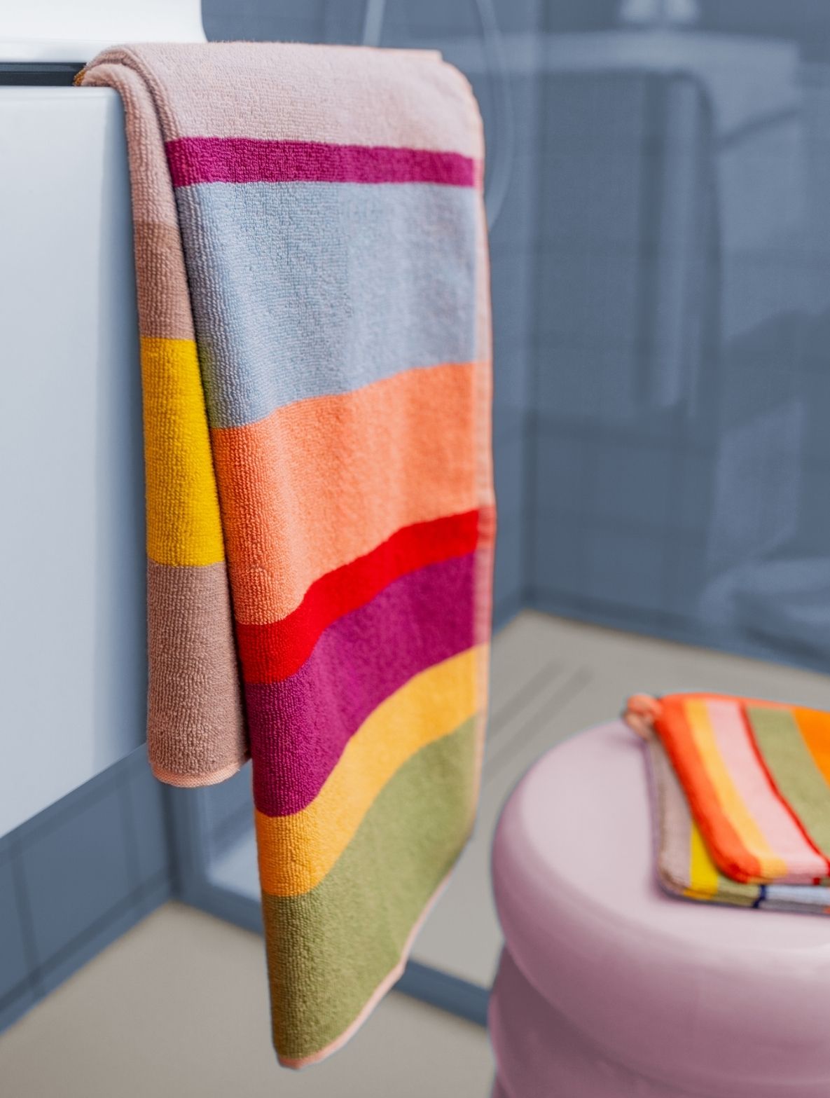 Ein großes, bunt gestreiftes Handtuch hängt über einem weißen Waschtisch im Bad. Es zeigt breite horizontale Streifen in leuchtenden Farben wie Gelb, Hellblau, Orange, Rot, Lila und Grün. Im Hintergrund: blaue Wand, rosa Hocker mit gefaltetem Handtuch.
