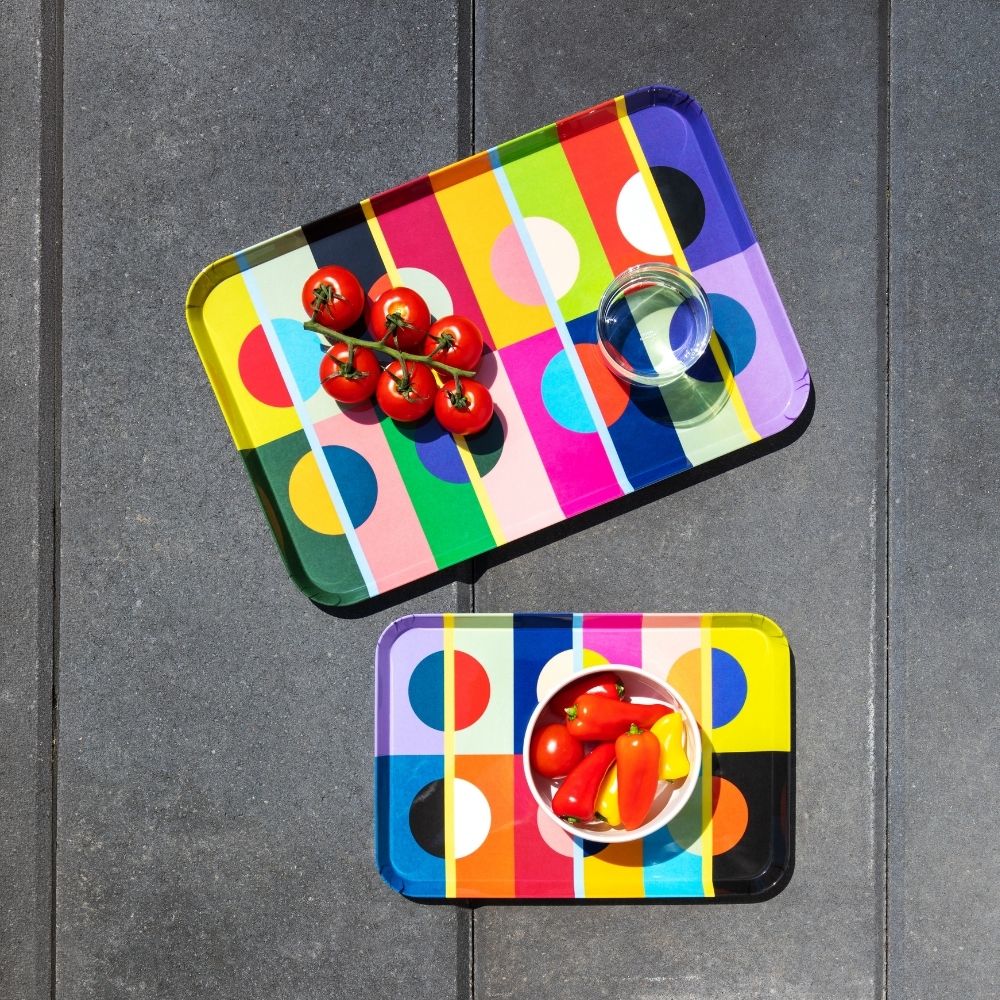 Zwei bunte, rechteckige Serviertabletts auf dunklen Terrassenfliesen. Oben: Rispe mit sechs roten Kirschtomaten und ein kleines Glas Wasser. Unten: weiße Schale mit roten und gelben Mini-Paprika. Geometrisches Muster.