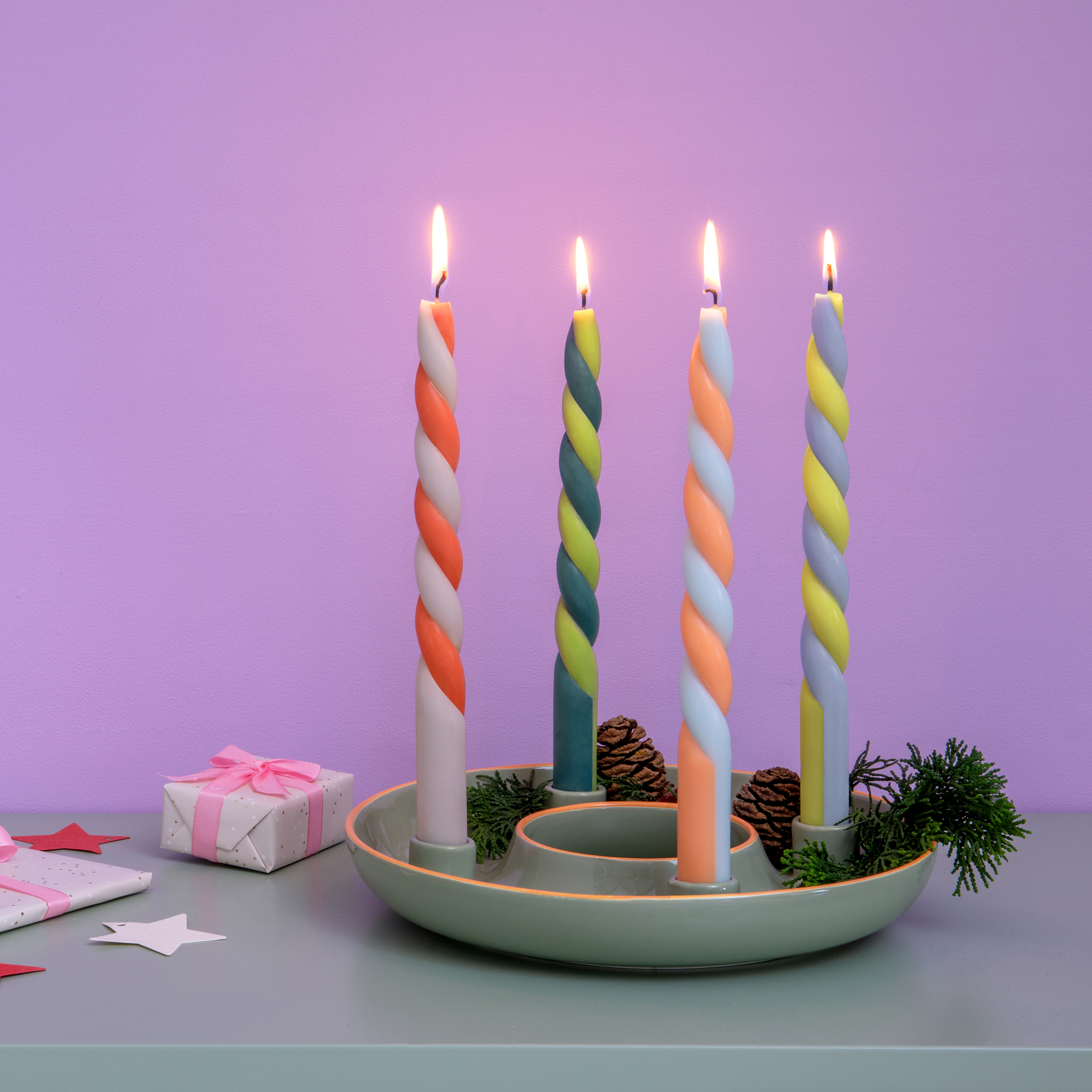 Velas cilíndricas «Helsinki» de colores, encendidas y retorcidas, en una bandeja verde con piñas y regalos sobre una mesa con fondo morado.