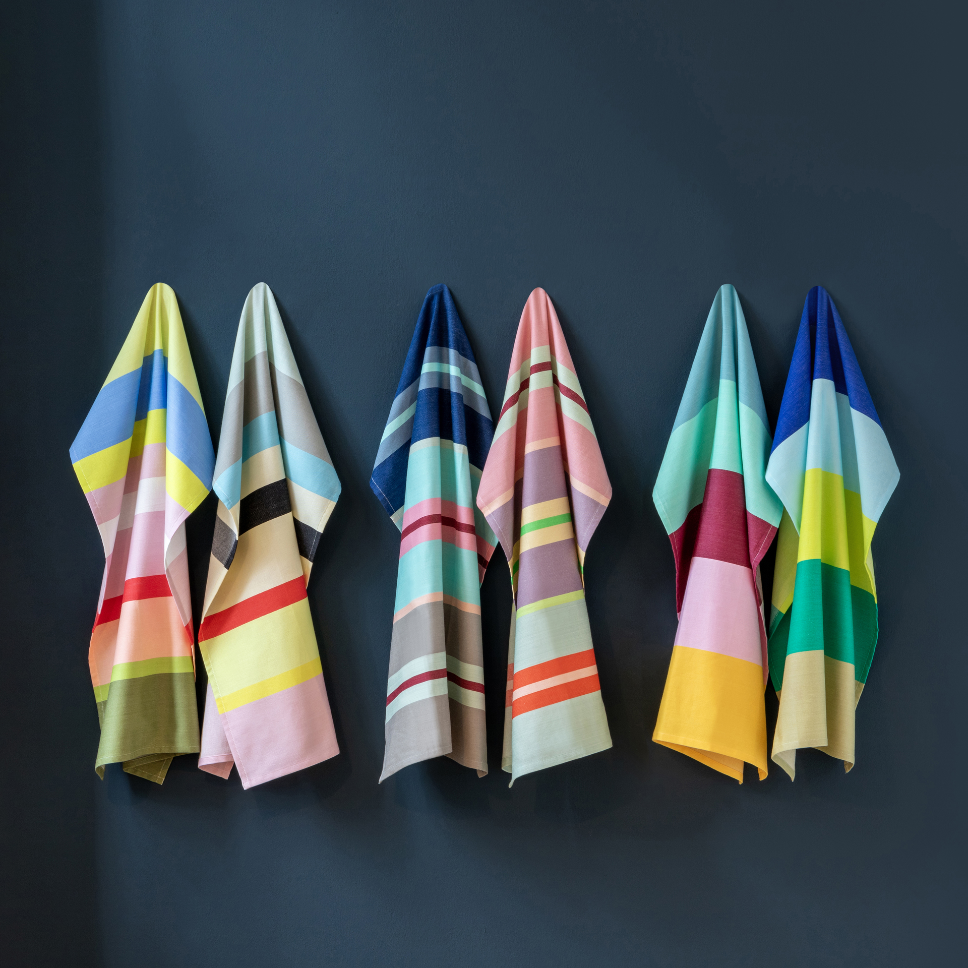 Six chiffons de séchage rayés Set n° 23 sont suspendus devant un mur sombre. Les torchons ont différentes combinaisons de couleurs, dont le jaune, le bleu, le rose et le vert.