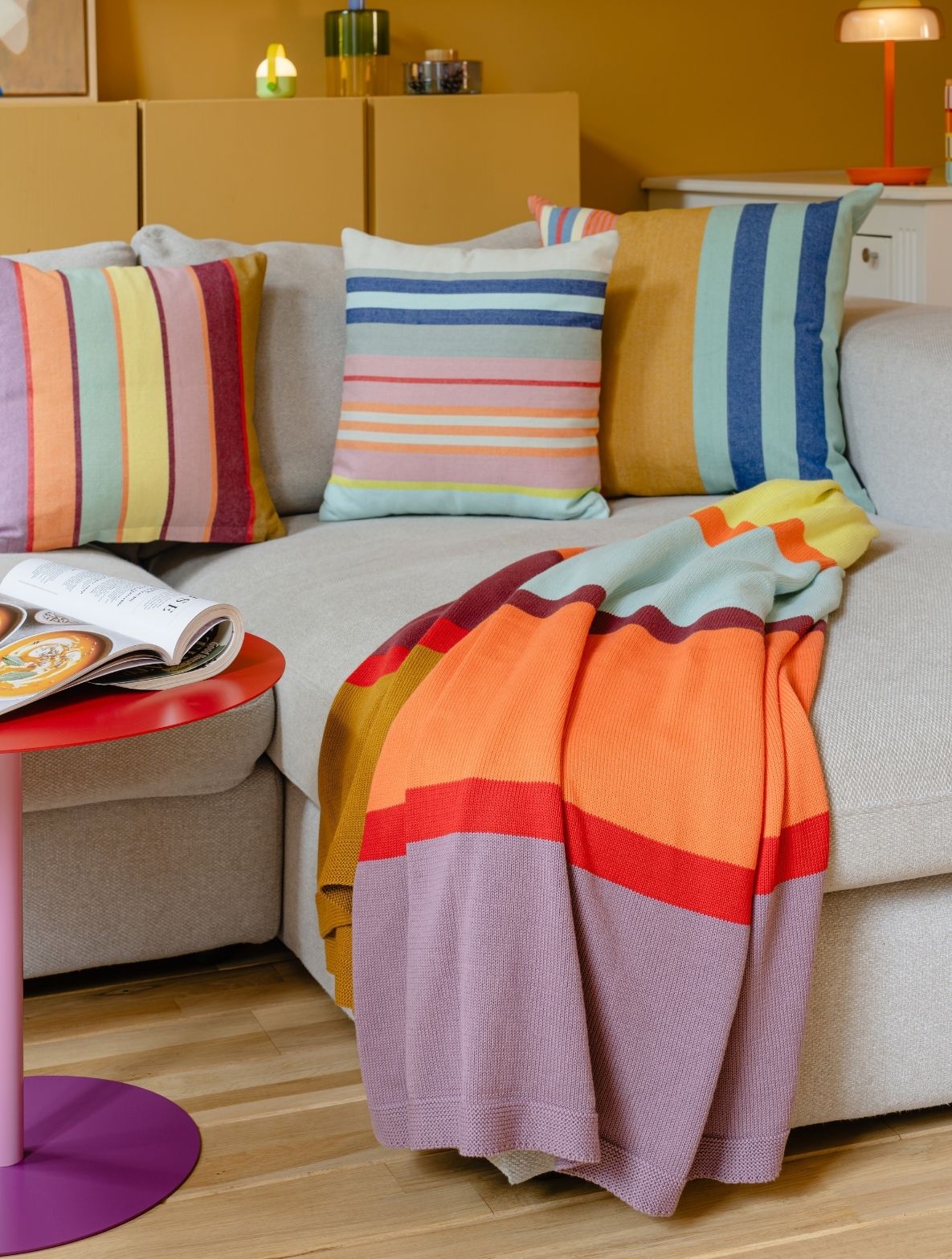 Beigefarbenes Sofa mit bunten Kissen in Streifenmustern und einer gestreiften Decke in Lila, Rot und Orange über der Armlehne. Links ein roter Beistelltisch mit offenem Magazin. Gemütlich, farbenfroh, vor senfgelber Wand.