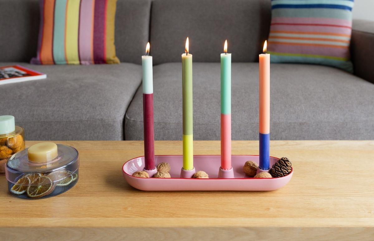 Vier brennende, zweifarbige Kerzen, oben hell, unten dunkel, in rosa Kerzenhalter auf Holztisch. Walnüsse und kleiner Kiefernzapfen zieren den Halter. Im Hintergrund graue Couch mit Streifenkissen und Dose mit Orangenscheiben.