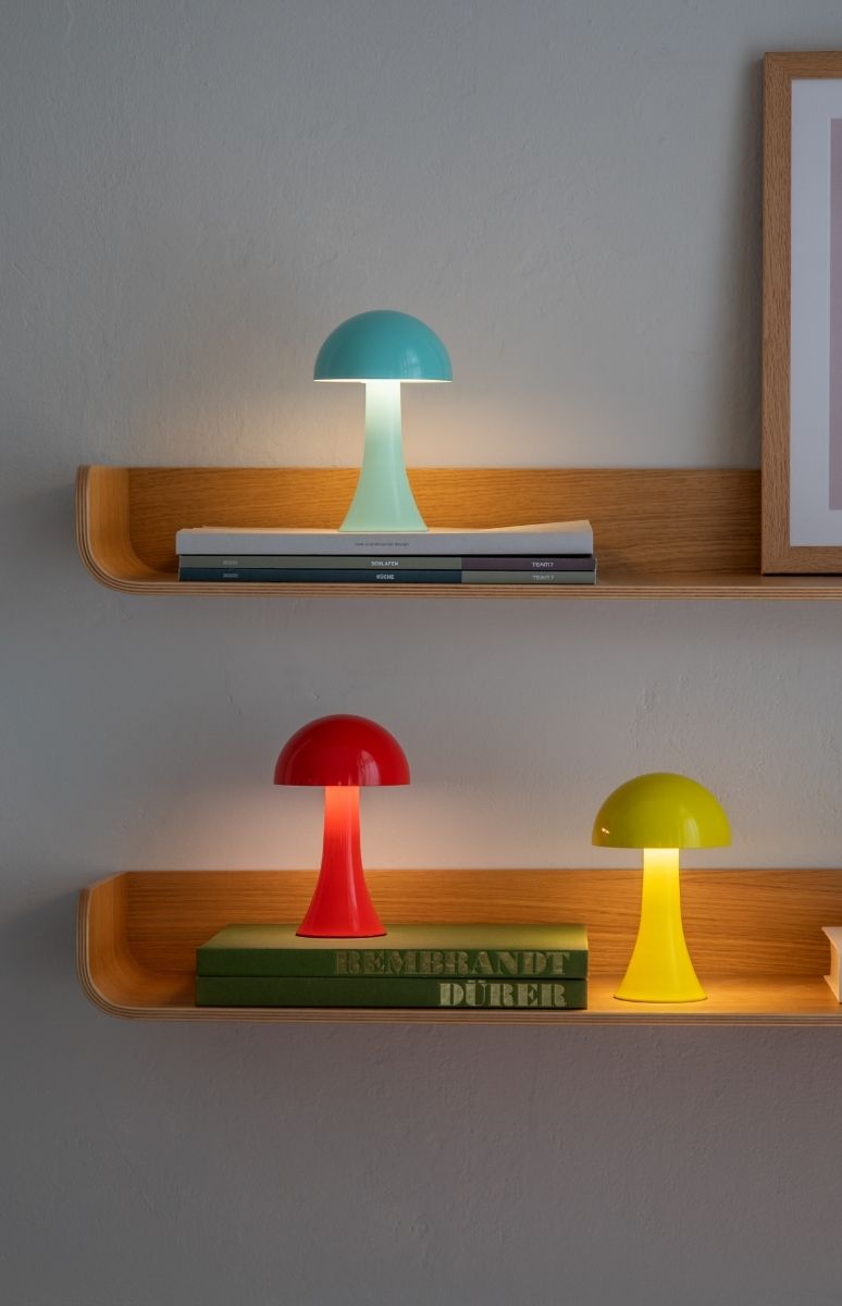 Drei moderne, pilzförmige Leuchten in kräftigen Farben auf zwei hellen Holzregalen vor grauer Wand. Oben eine türkisfarbene Lampe, unten links rot und rechts gelb; alle beleuchten Bücher mit warmem Licht nach unten.
