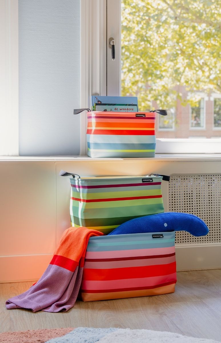 Drei bunte, horizontal gestreifte Aufbewahrungskörbe stehen neben einem Fenster übereinander. Im obersten Korb auf der Fensterbank liegen Bücher. Unten sind eine orange Decke und ein blaues Wal-Stofftier. Draußen ist grünes Laub zu sehen.