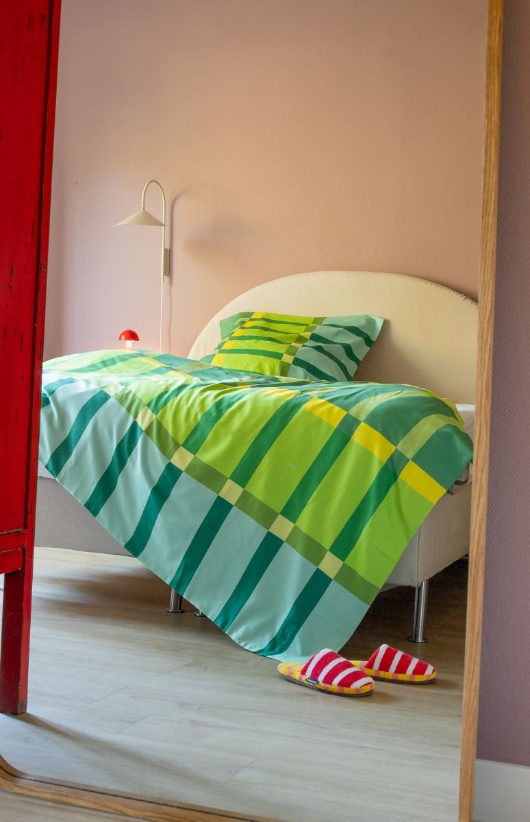 Ein gemütliches Schlafzimmer mit Bett, bezogen mit Bettwäsche in Grün-, Hellblau- und Gelbtönen mit geometrischem Muster. An der rosa Wand hängt eine weiße Leselampe. Rot-weiß gestreifte Hausschuhe liegen auf Holzboden.