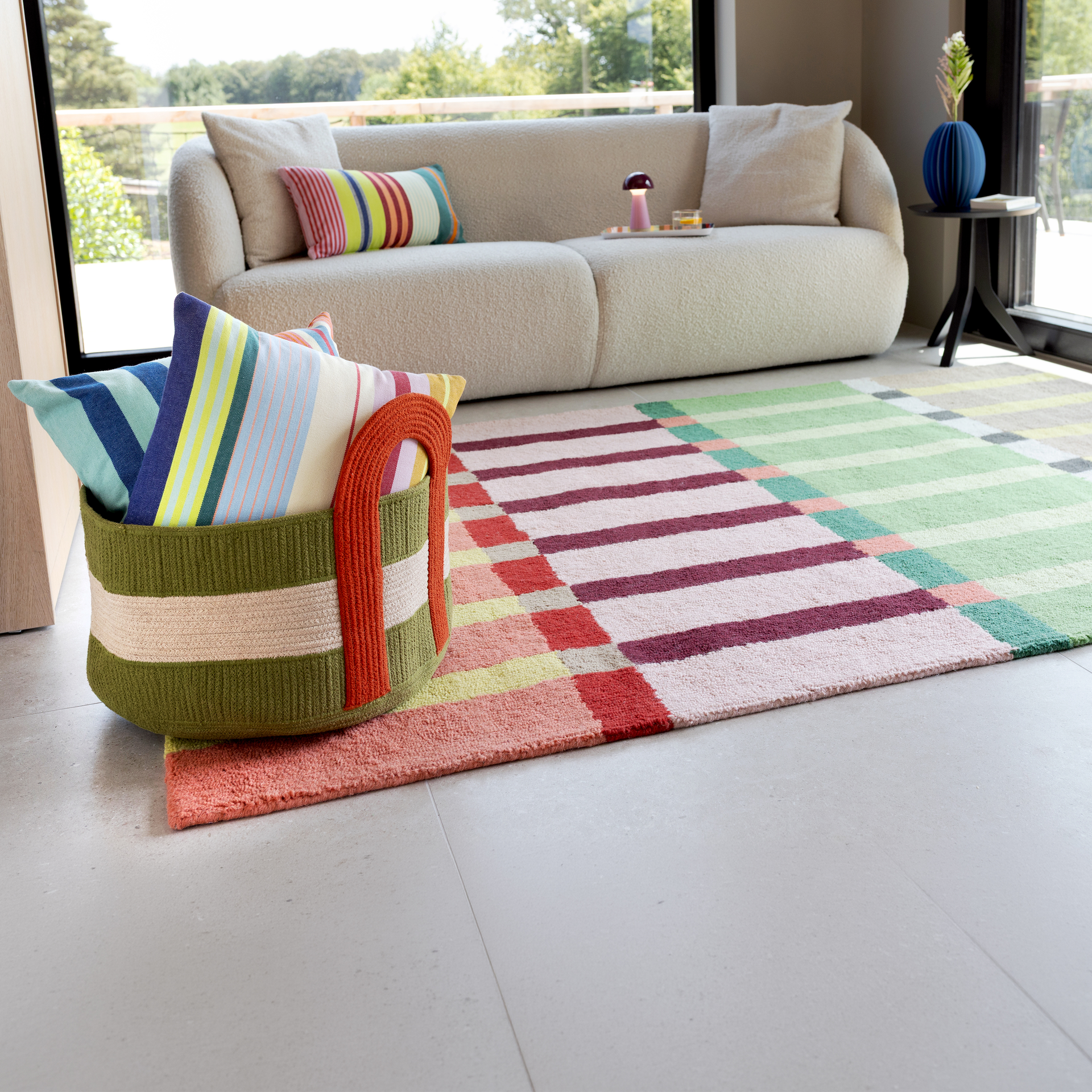 The ‘Kaido’ rug, with eye-catching striped and block patterns in vibrant colours like pink, red, green and yellow, stands out in the modern living room. It lies on a light-coloured tiled floor. Next to it is a woven basket of cushions.